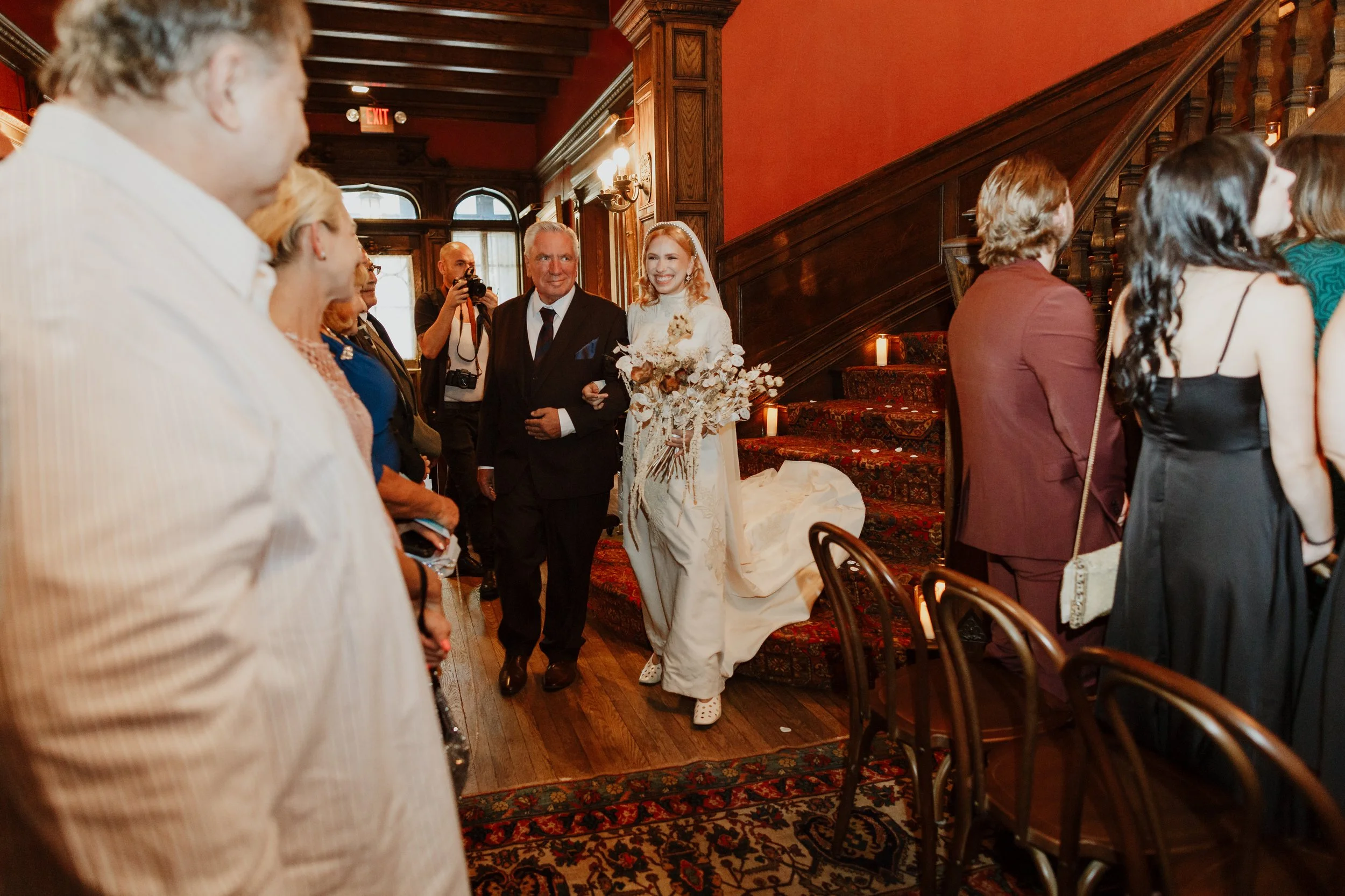 A bride in a white wedding dress holding a bouquet of white flowers walking down a staircase at a wedding reception, smiling and facing a man in a dark suit. Guests are seated and standing along a wooden staircase, watching her. The room has warm lig