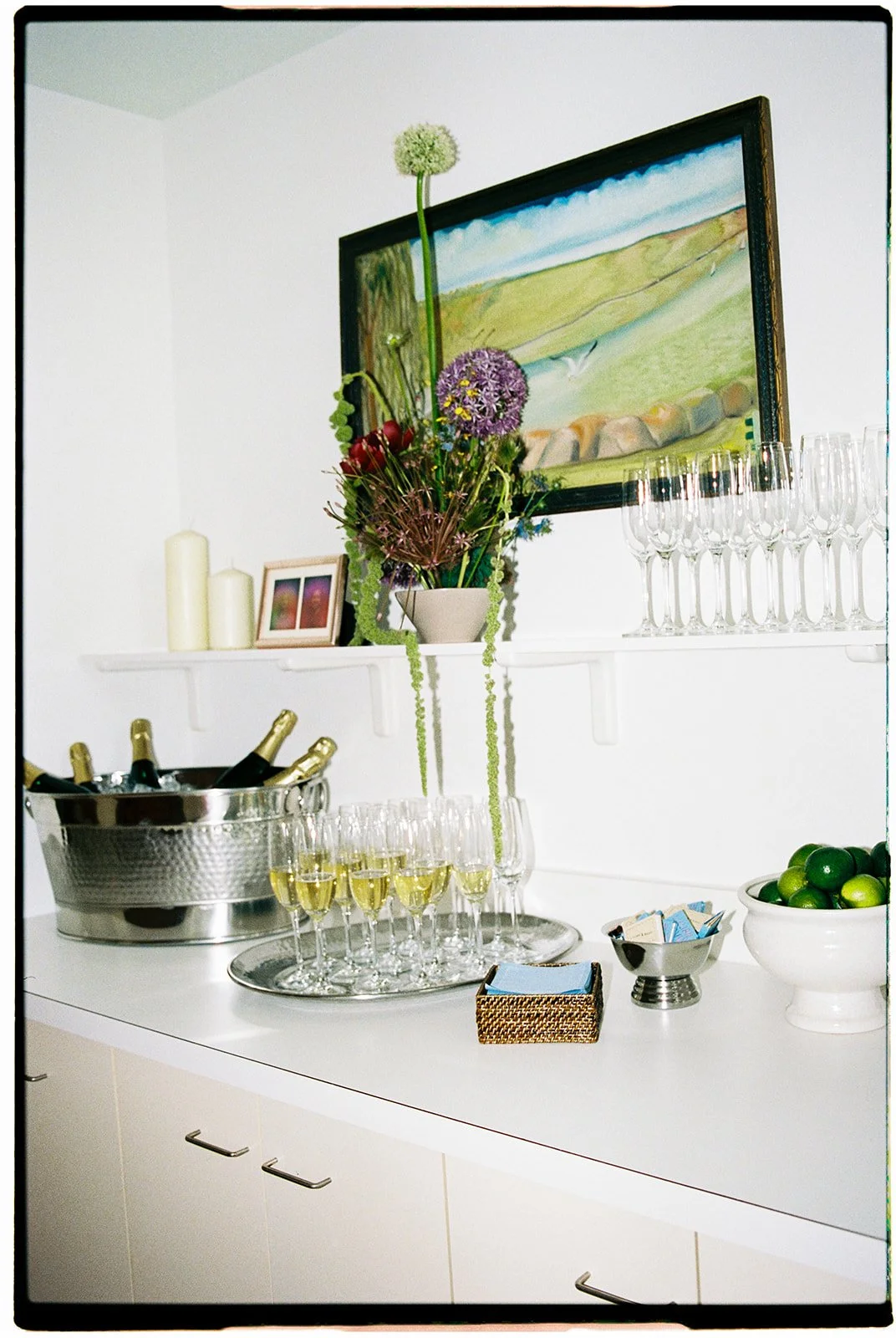 A white table with a silver champagne bucket containing bottles of champagne, a tray of champagne glasses filled with bubbly, a bowl of green limes, and a small basket of sugar packets. A floral arrangement with tall green, purple, and red flowers is