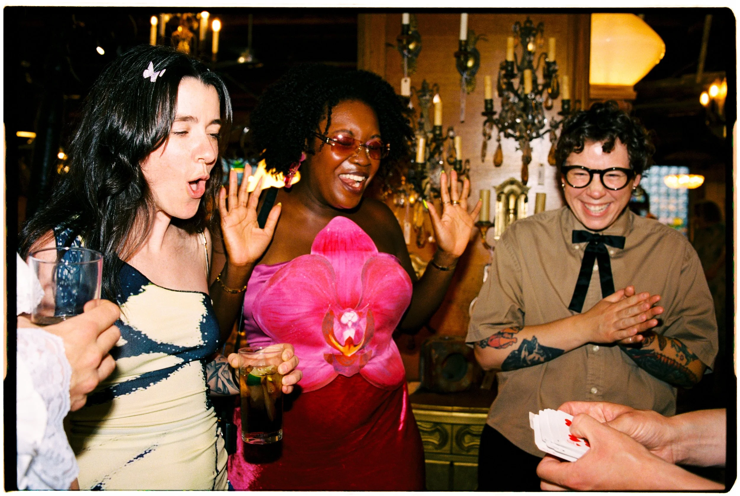 Group of women at a social gathering playing a card game, smiling and enjoying themselves, in a warmly lit room with candles and ornate decorations.