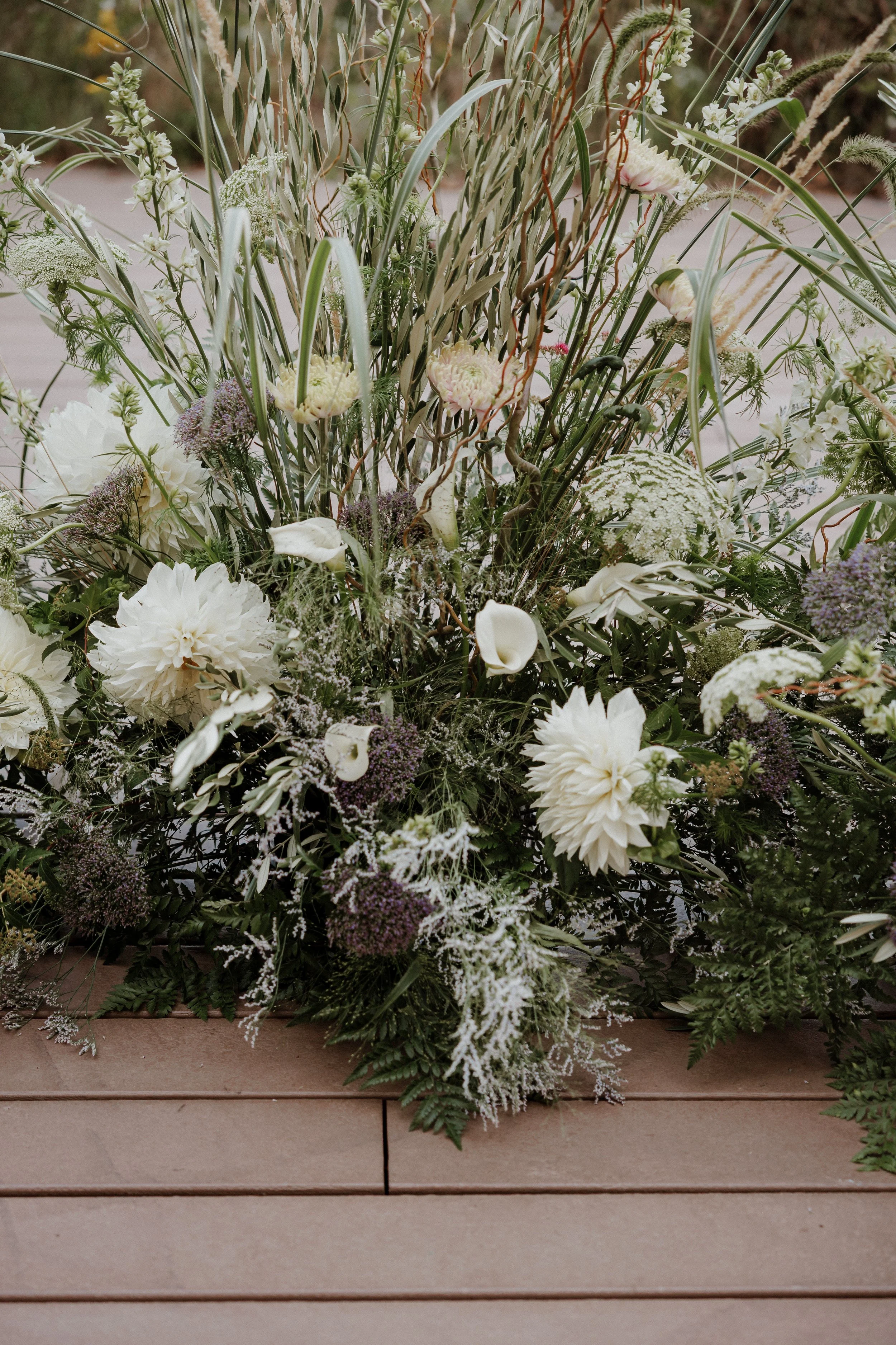 A large floral arrangement with white and purple flowers, greenery, and grasses on a wooden surface.