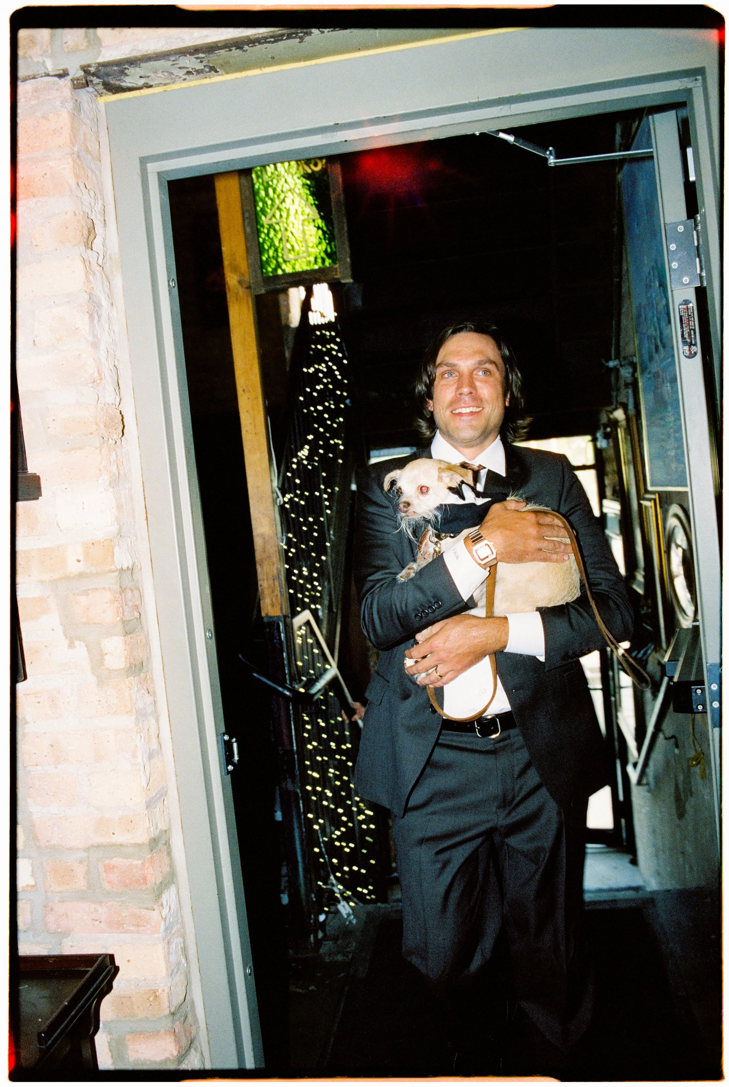A man in a black suit holding a small dog while smiling, standing in a doorway with exposed brick and industrial decor in the background.