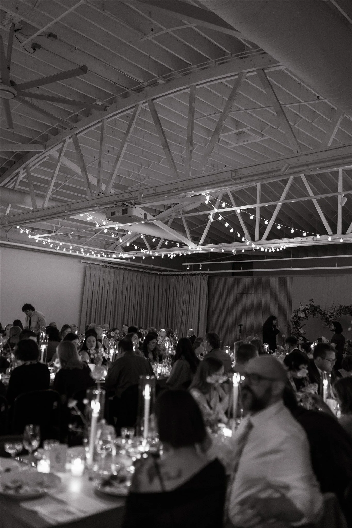A large group of people seated at round tables decorated with floral centerpieces and lit by candles, in a spacious hall with a high ceiling, string lights, and exposed beams. Some people are eating, and a woman is standing at the front near a floral