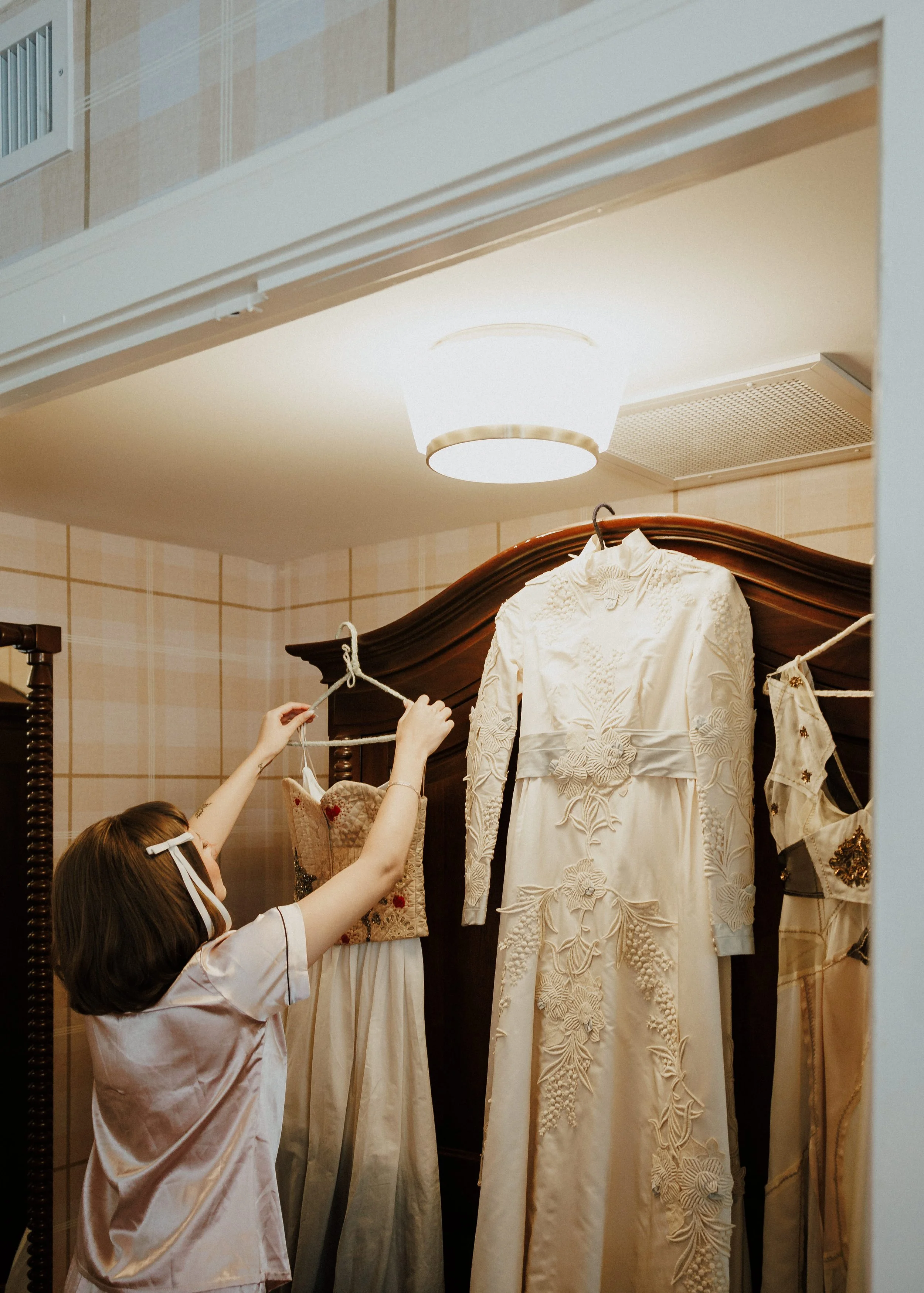 A woman, wearing a pink satin robe, is hanging up wedding dresses on a clothing line inside a fitting room or bridal boutique.