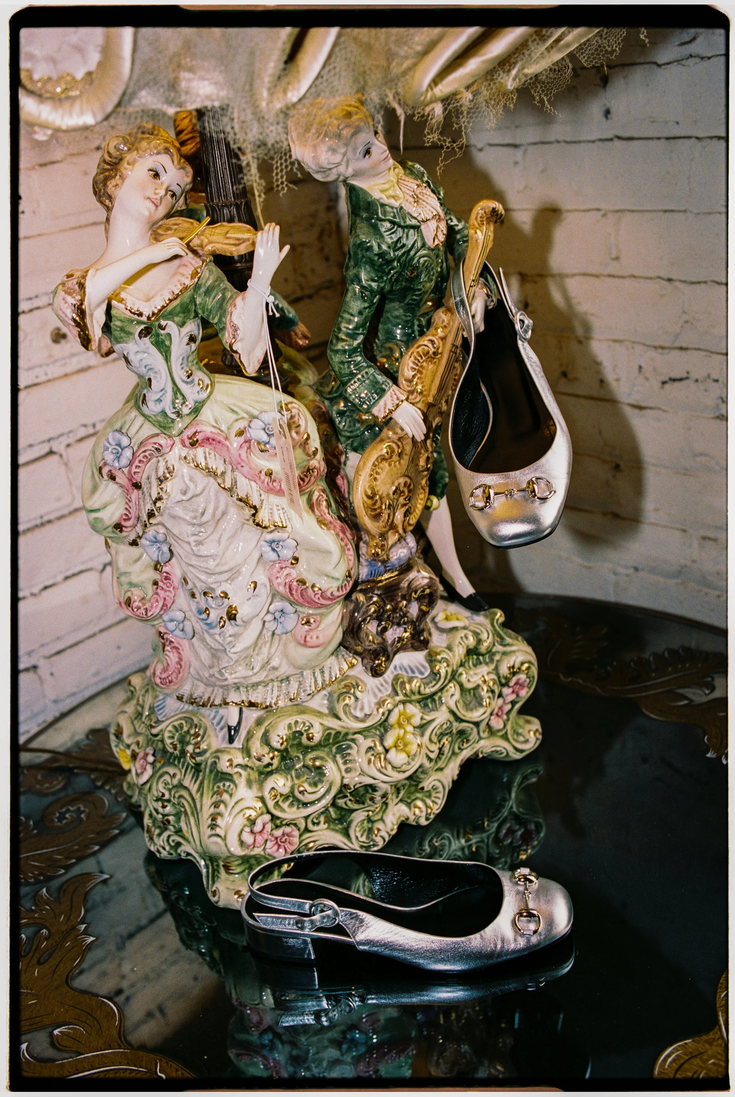 A porcelain figurine of a woman in a colorful, ornate dress playing the violin, with a young man in a green and gold jacket holding a musical instrument. The figurine has intricate detailing and is situated on an ornate, floral-patterned base. A meta