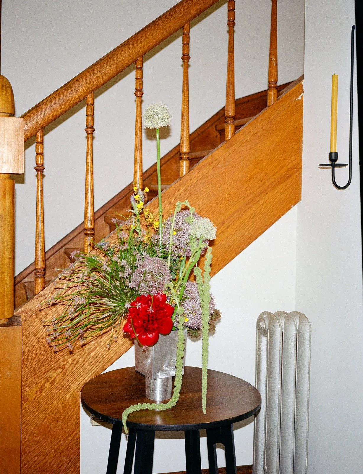 A wooden staircase with vertical spindles and a curved handrail, decorated with a floral arrangement in a silver vase on a small black side table near a white wall with a black and yellow wall sconce and a radiator.