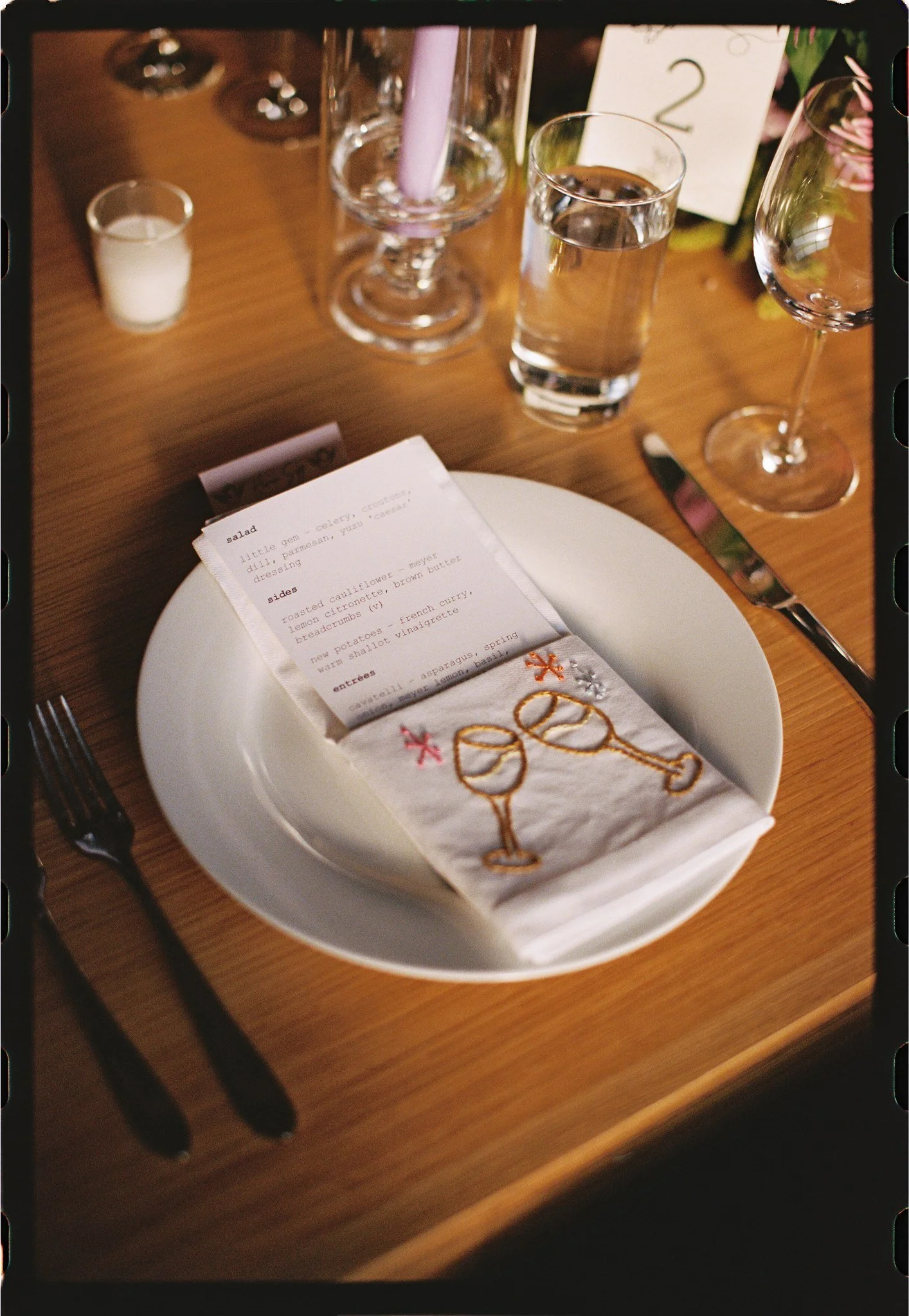 A table setting with a white plate holding a paper napkin embroidered with champagne glasses, a printed menu, a fork on the left, and a knife on the right. In the background, there are two wine glasses, a glass of water, a small candle, and a candle 