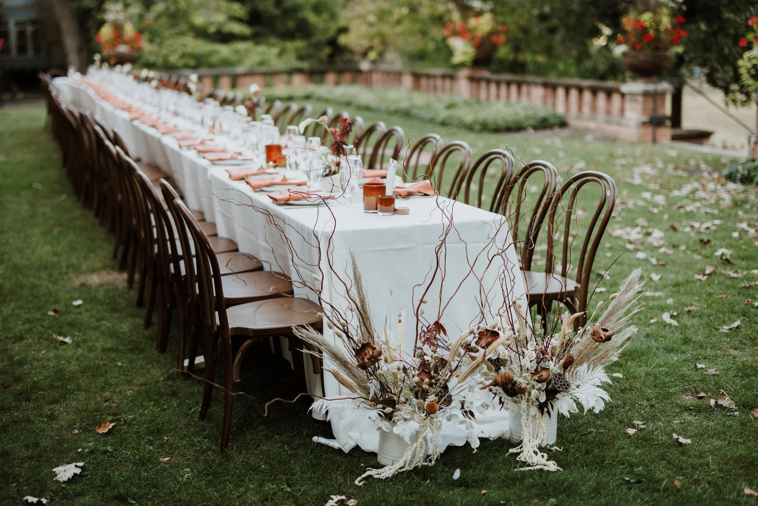 Long banquet table set outdoors on grass with white tablecloth and place settings, decorated with a large arrangement of dried flowers and foliage in front.