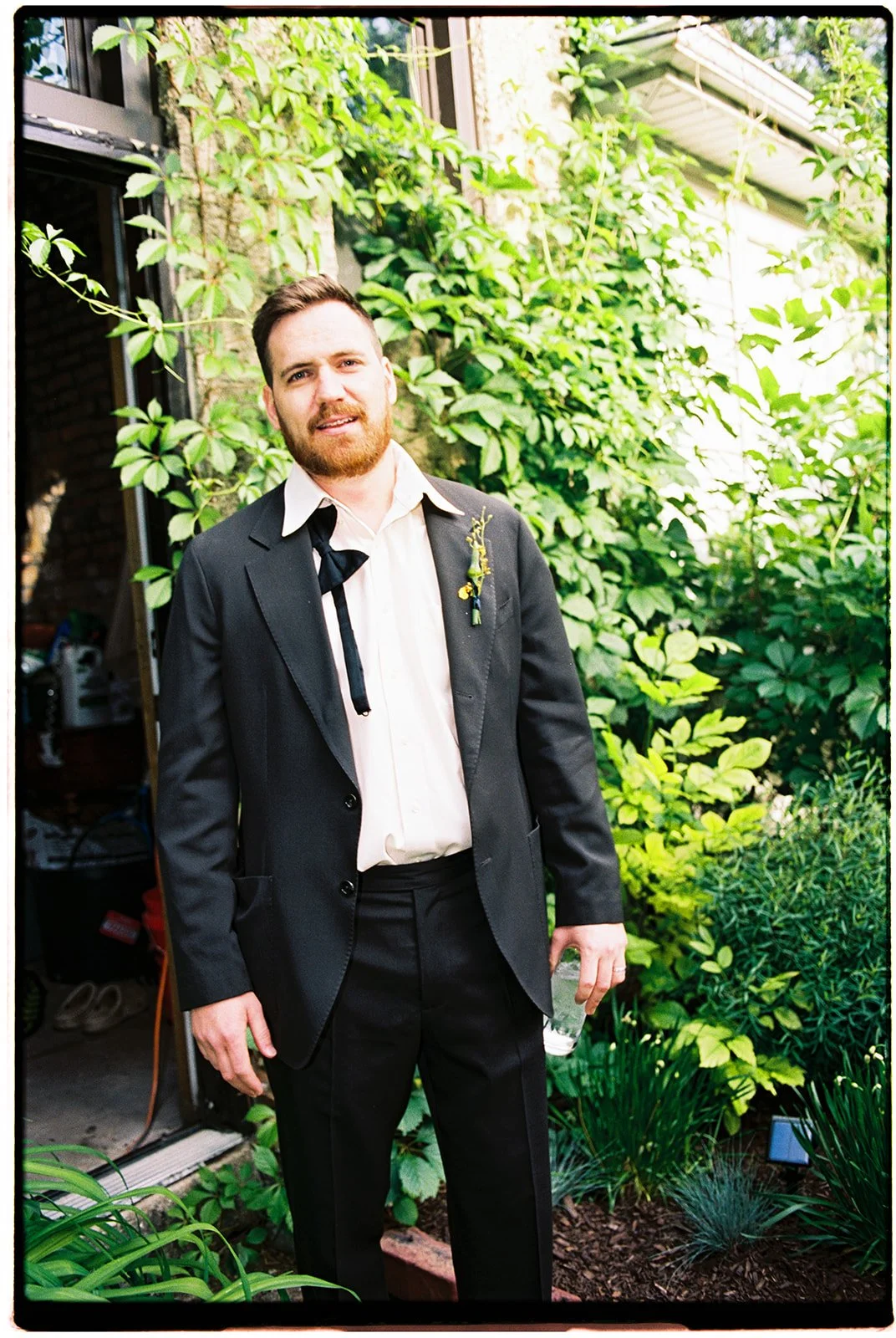 A man in a black suit with a white shirt and black tie standing in a lush green garden, holding a glass of water, with vines and plants in the background.