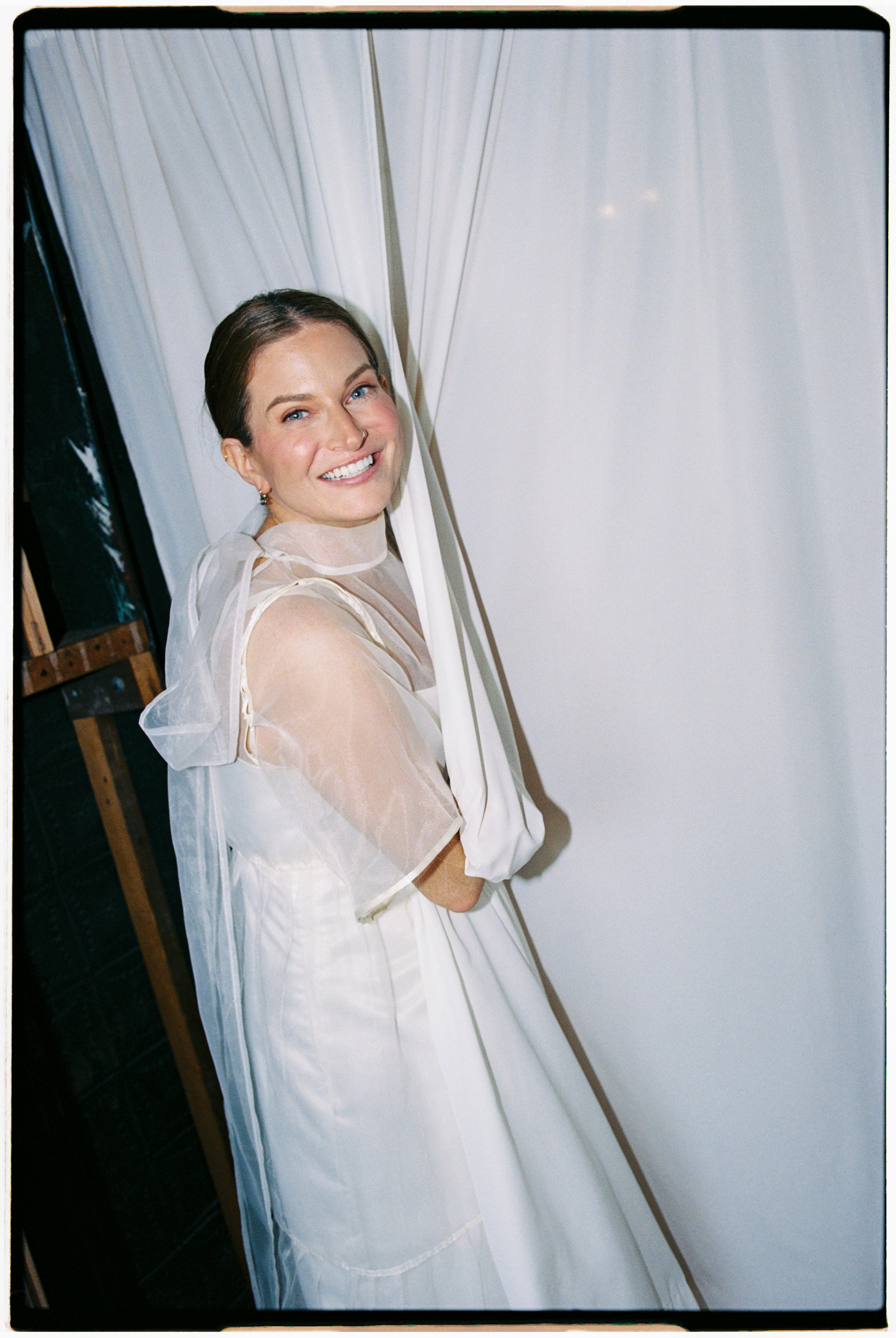 A woman with medium-length brown hair, blue eyes, and a nose ring smiling, dressed in light-colored, semi-sheer fabric, standing behind semi-transparent curtains in a brightly lit room.