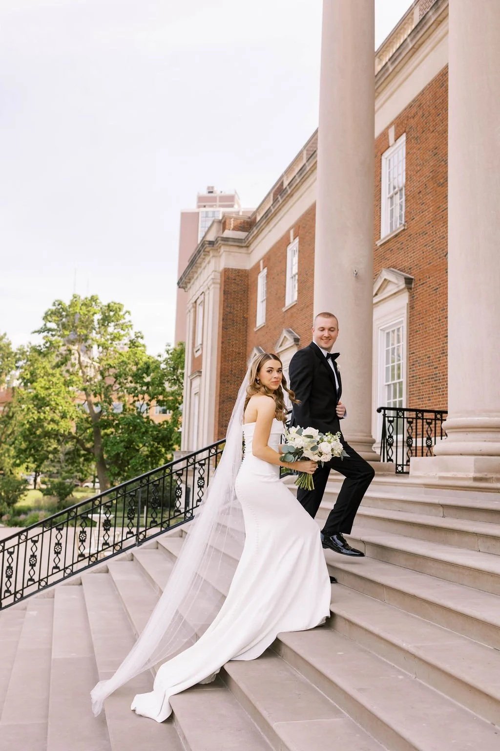A bride and groom standing on the steps of a historic building with large columns, holding a bouquet of white flowers, dressed in wedding attire.