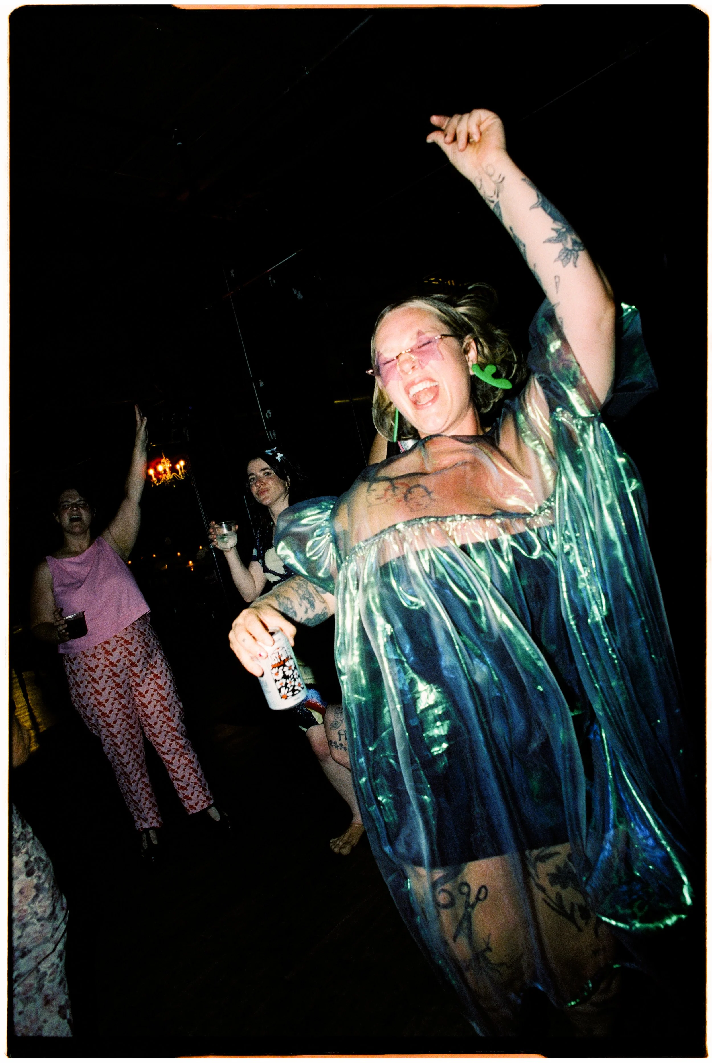 A woman with tattoos wearing a shiny, iridescent, short-sleeved dress, pink glasses, and colorful earrings, dancing and singing at a party or celebration, holding a bottle of beer, with a joyful expression on her face. Other women are seen in the bac
