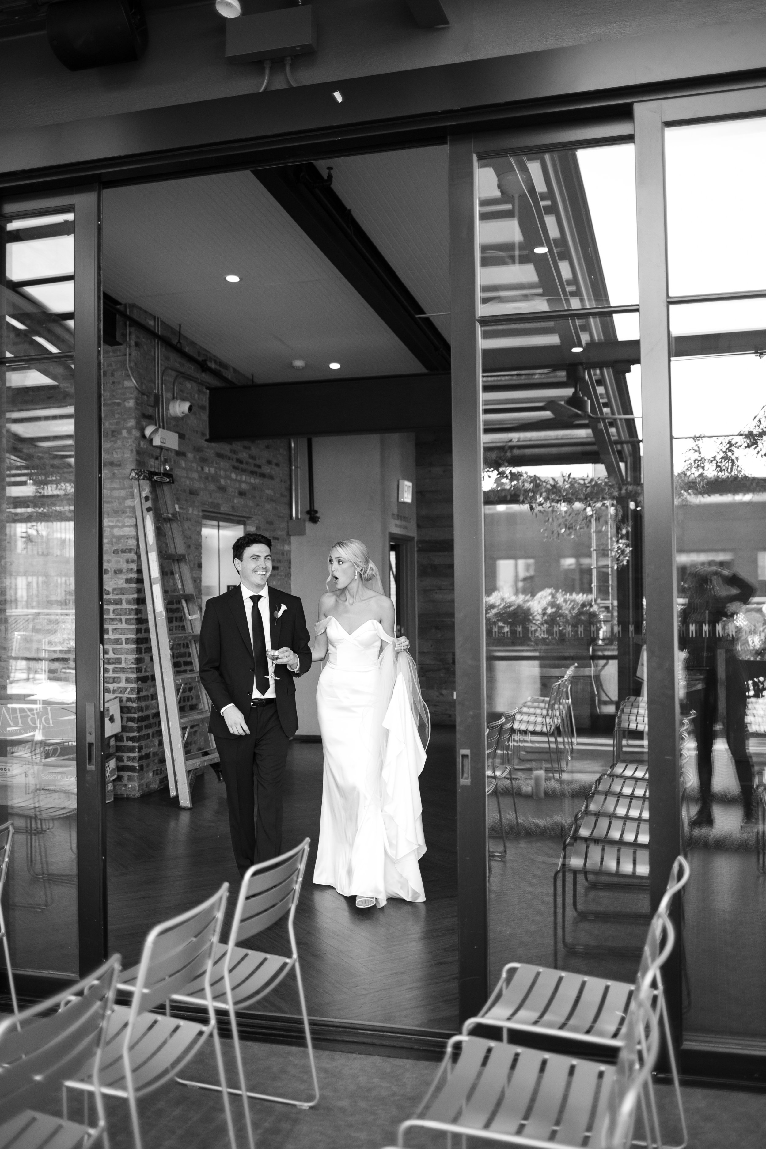 A bride and groom walking inside a modern venue with large glass windows, sitting chairs in the foreground, brick and wood interior, and outdoor greenery visible.