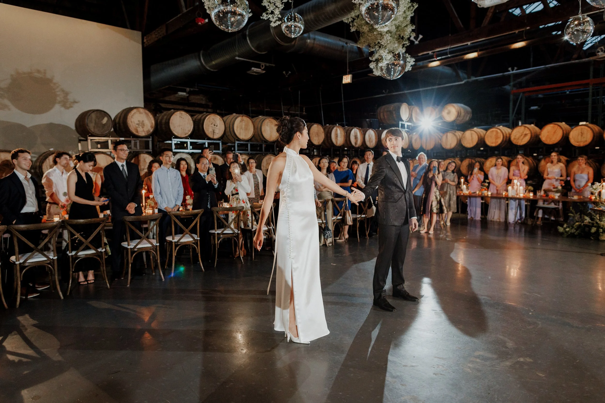 A bride and groom holding hands during their wedding reception in a rustic venue with wine barrels and guests in elegant attire watching.
