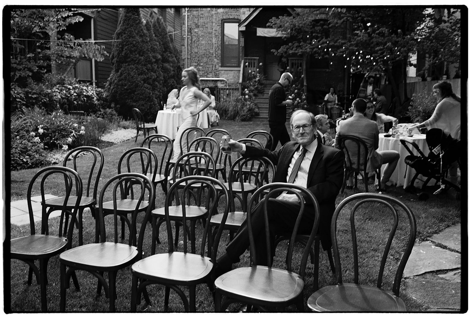 People gather for a backyard celebration, with some sitting at tables and others standing or walking around, surrounded by trees and string lights.