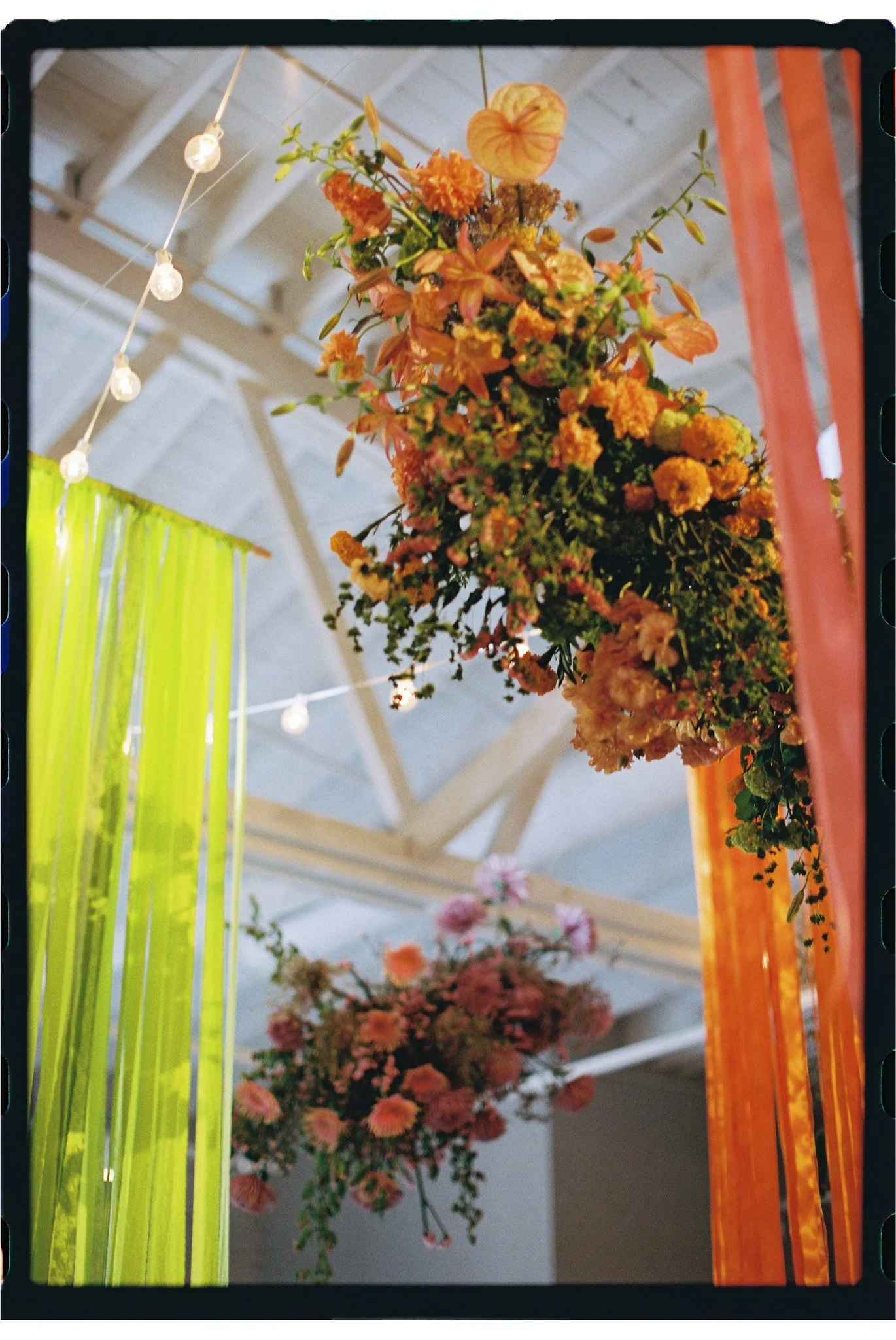 Hanging floral arrangement with orange and yellow flowers, and greenery, in an indoor space with string lights and colorful vertical panels in the background.