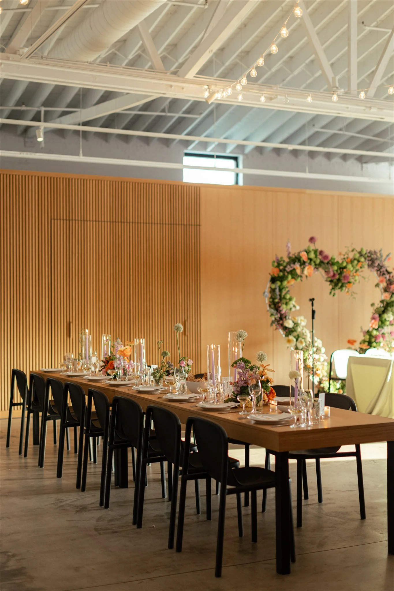 Indoor event space decorated for a celebration with a long banquet table set with floral centerpieces, candles, and place settings, featuring a wooden wall, large arch of flowers, and string lights overhead.