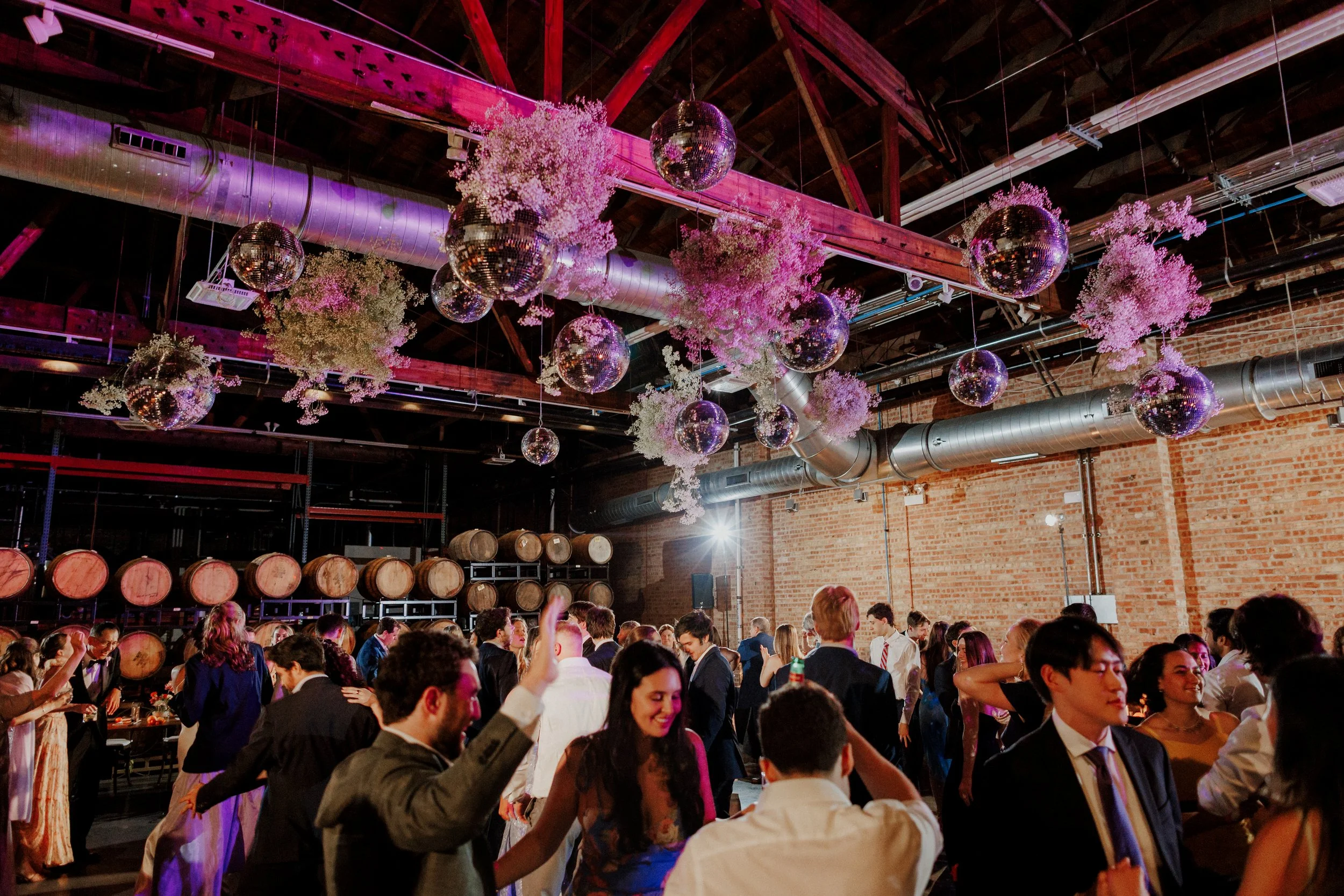 People dancing at a lively indoor party with disco balls, pink and purple lighting, and wine barrels against brick walls.
