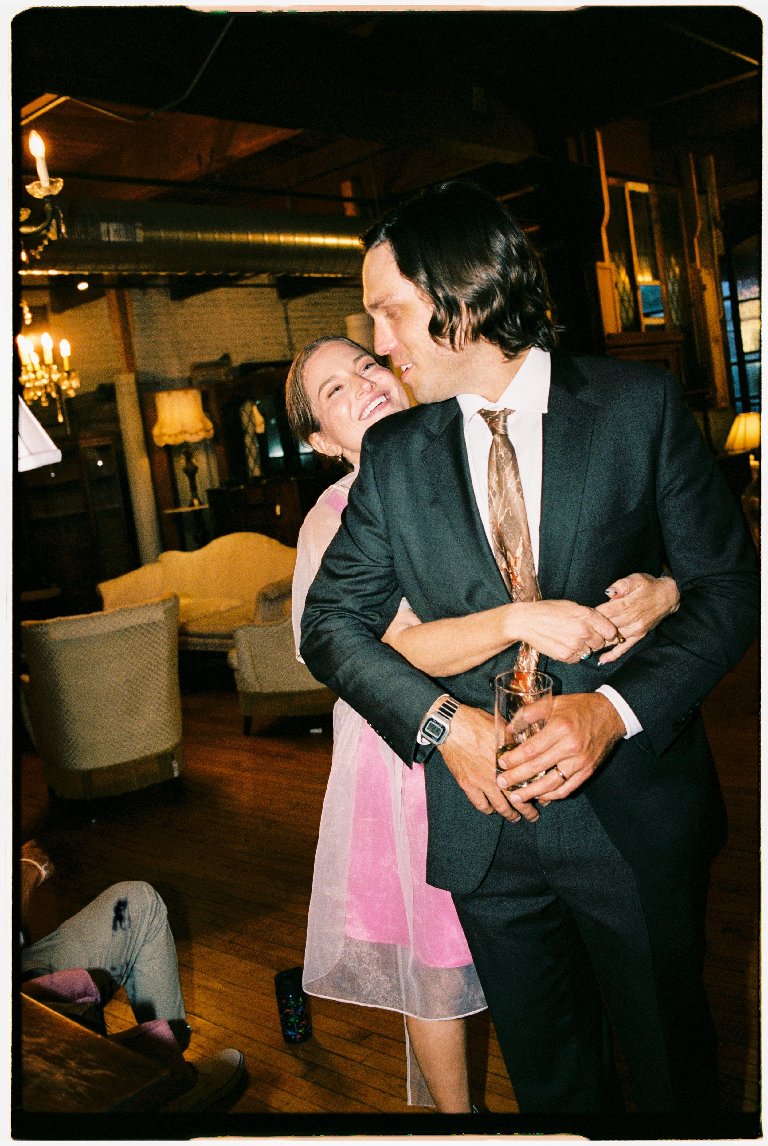A woman and a man sharing a close moment, smiling and embracing each other in a cozy, warmly lit restaurant or bar. The woman has light skin and is wearing a sheer pink and white dress, while the man has light skin, dark hair, and is dressed in a bla