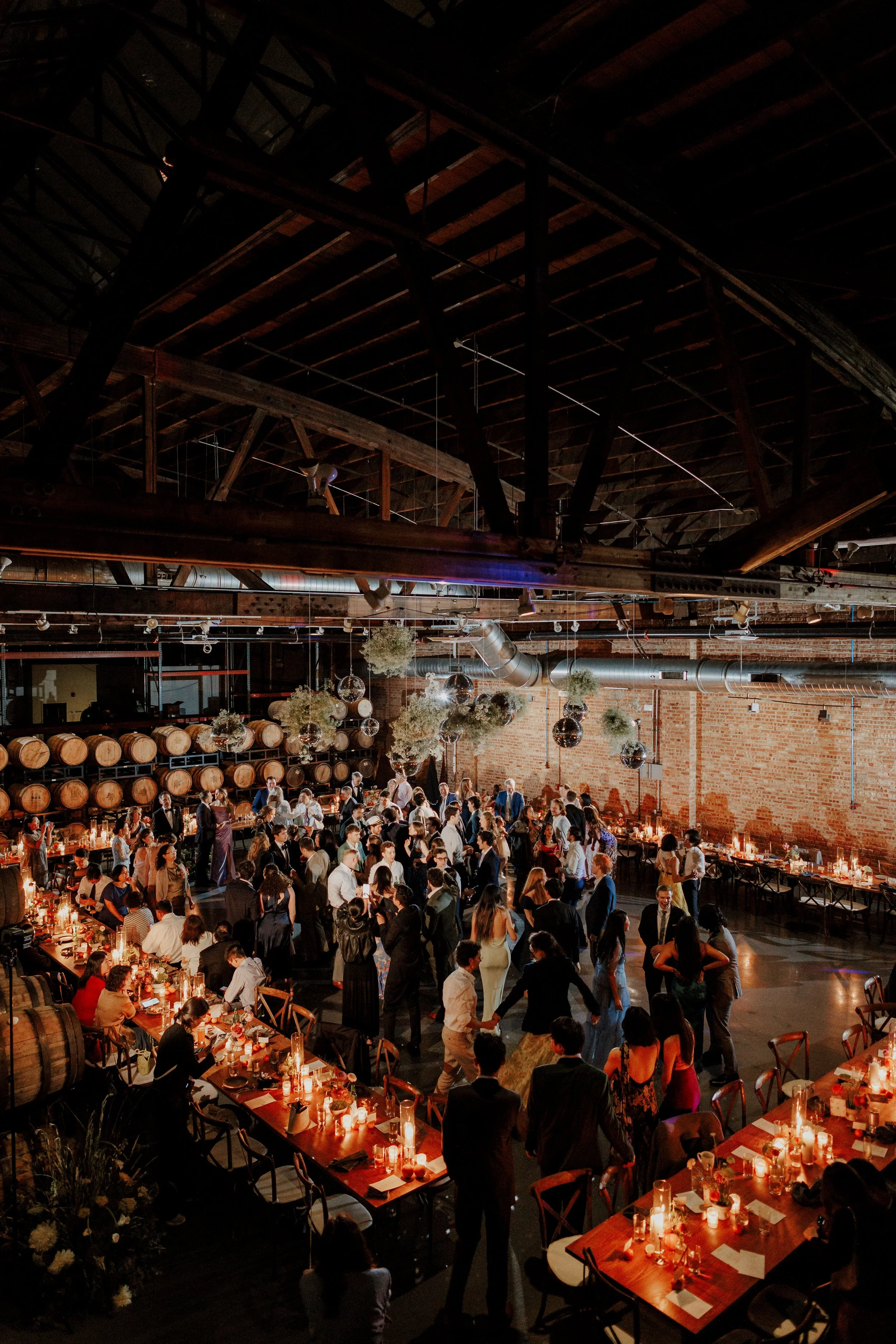 A large indoor event with many people socializing and dancing. The room has industrial decor with exposed brick walls, hanging plants, and vintage wine barrels. There are round tables with candles and place settings, and the ceiling has exposed beams