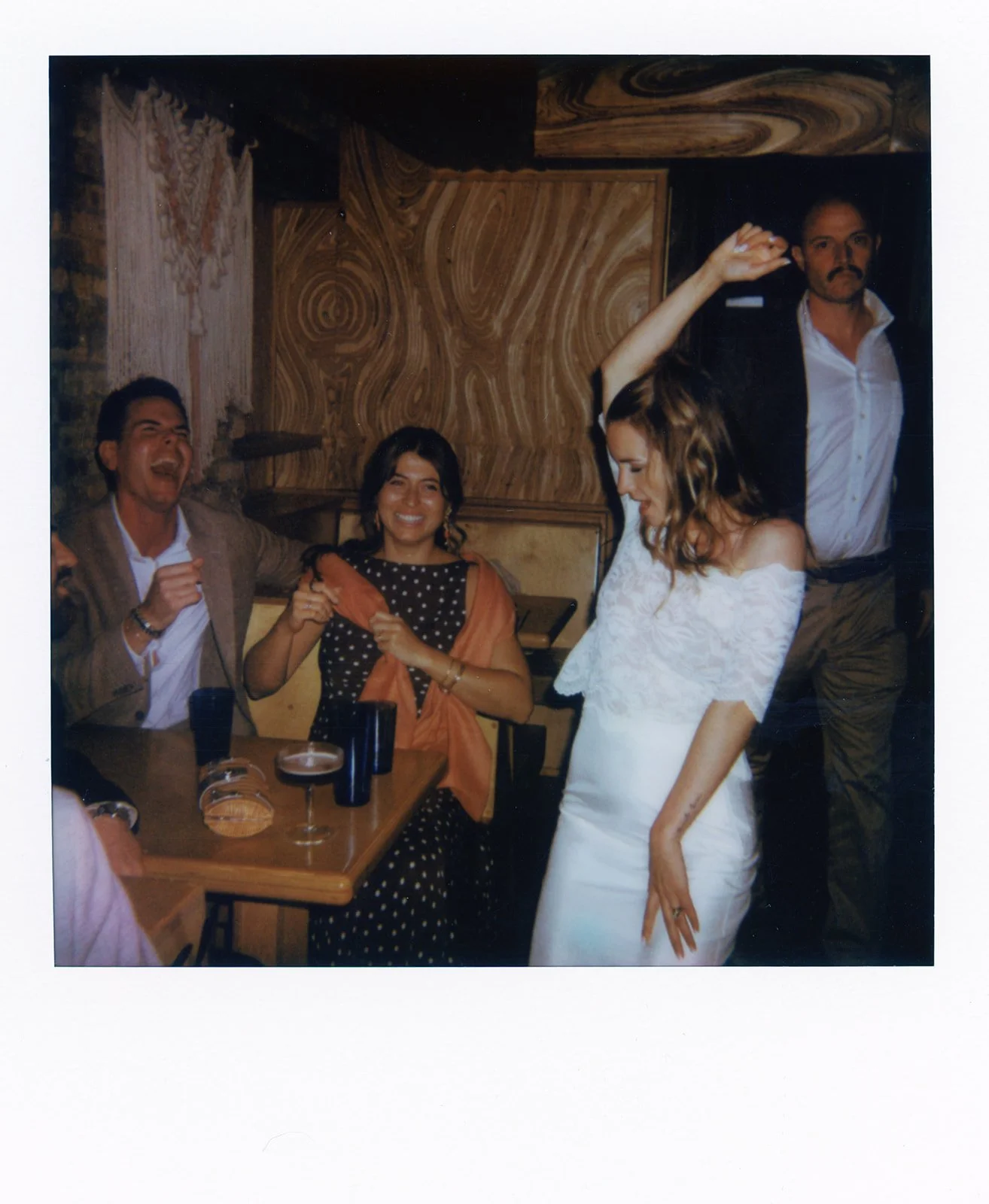 Group of four people enjoying a celebration in a cozy, wood-paneled restaurant. One woman in a white dress is dancing, while the others are sitting at a table laughing and smiling.