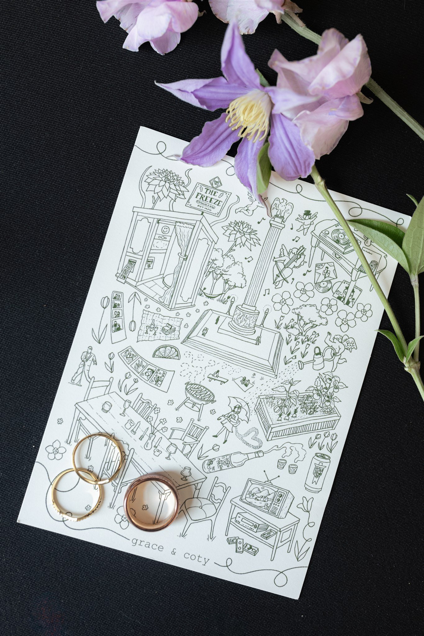 A coloring sheet with various illustrations of household items and decorative objects, placed on a black surface. Resting on top of the sheet are gold and rose gold rings, while a light purple and white flower with green leaves is partly covering the