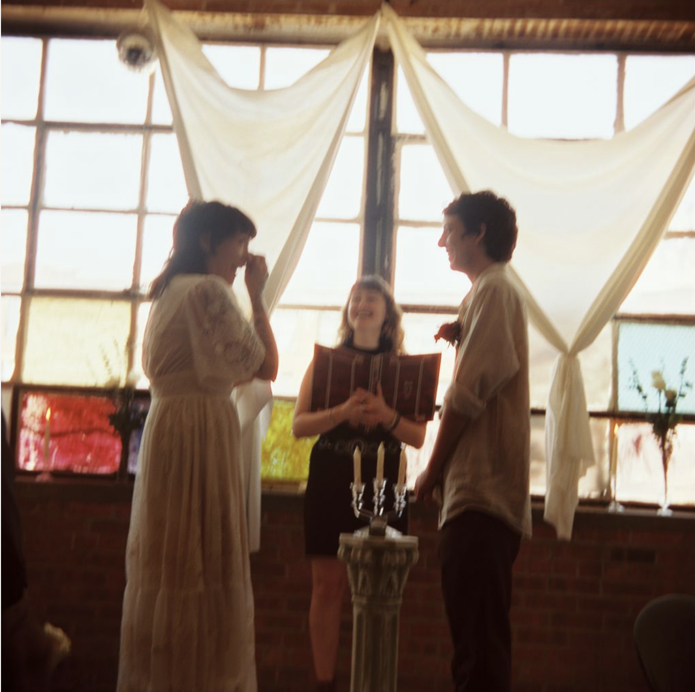 A couple getting married during an indoor ceremony with an officiant holding a book, in front of large windows with white curtains, and a candle on a pedestal.