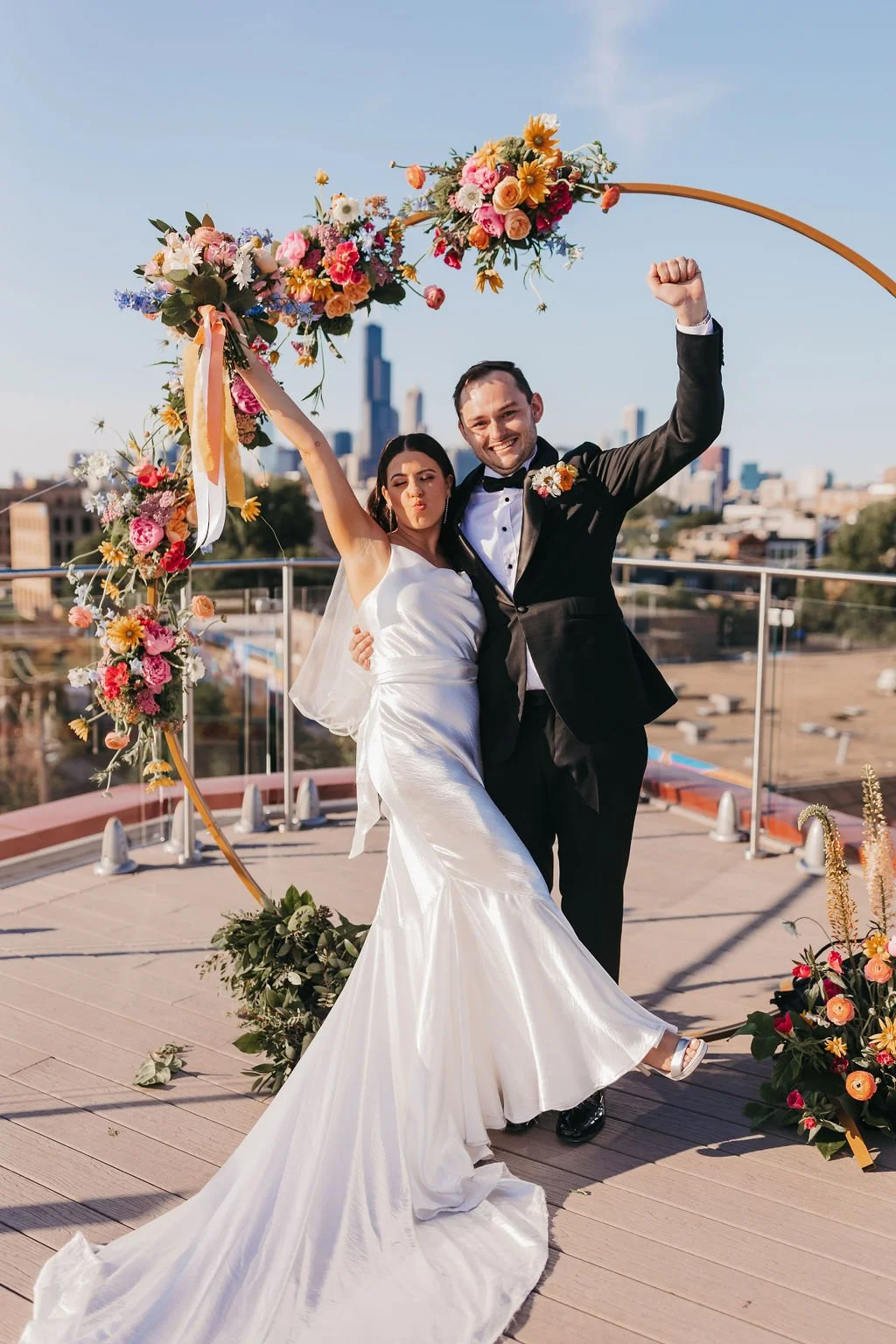 A newlywed couple in wedding attire celebrating on a rooftop with a city skyline in the background, standing under a floral arch with colorful flowers.