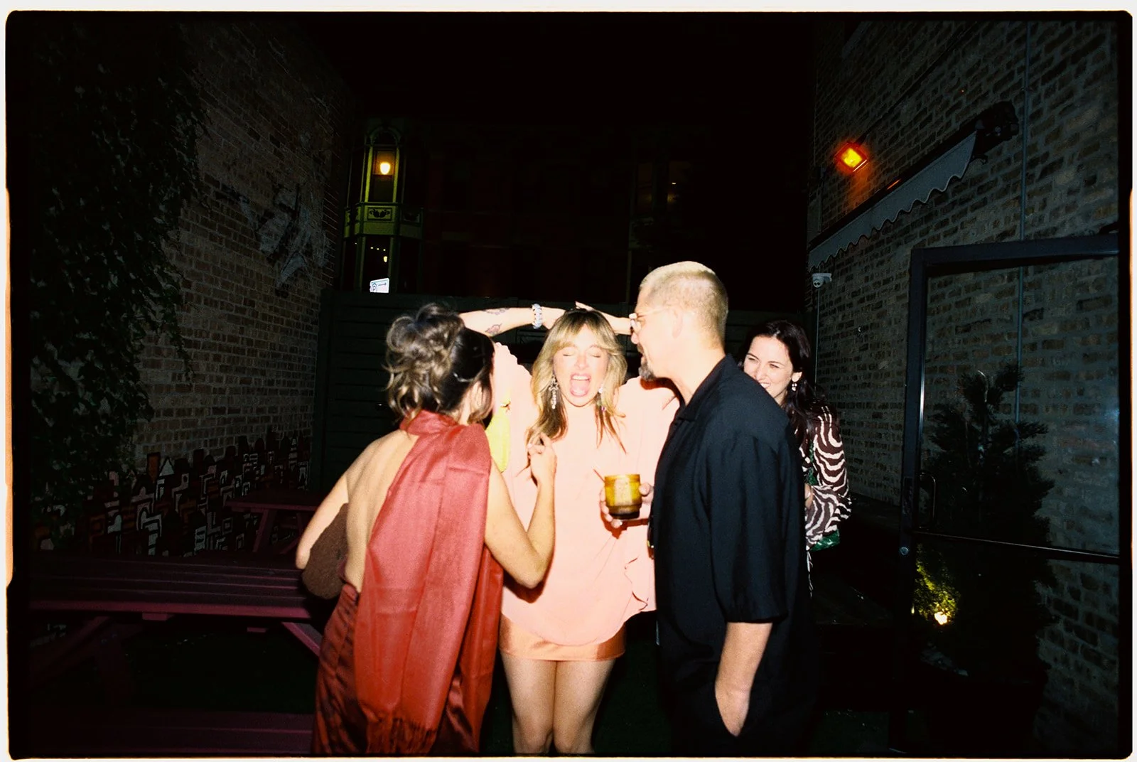 Group of five friends laughing and talking on a rooftop patio at night, with brick walls and greenery, one holding a drink.