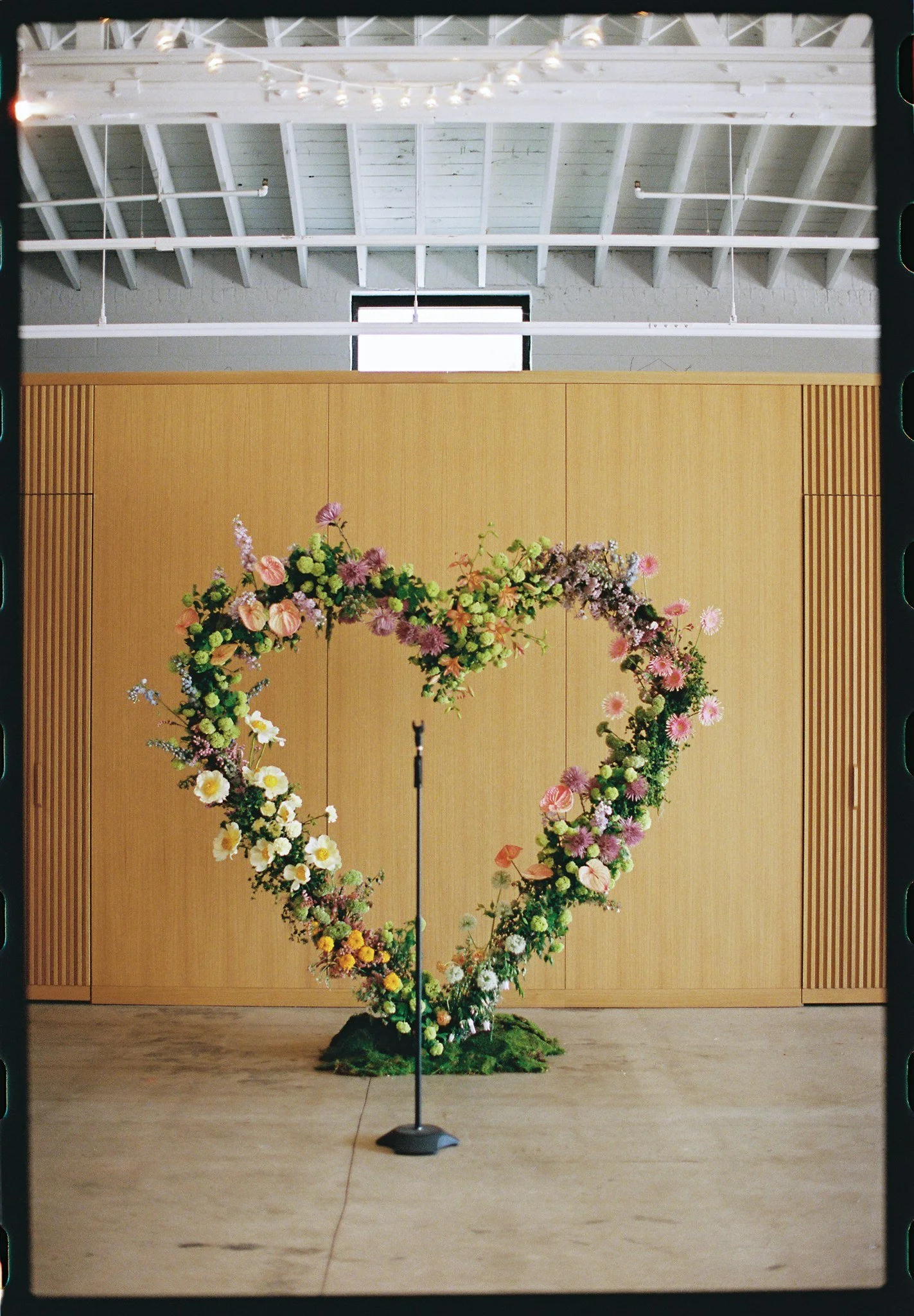 A floral wreath made of various pink, white, and purple flowers with green foliage, layered in a semi-circular shape, standing on a black base in front of a wooden wall with vertical slats.