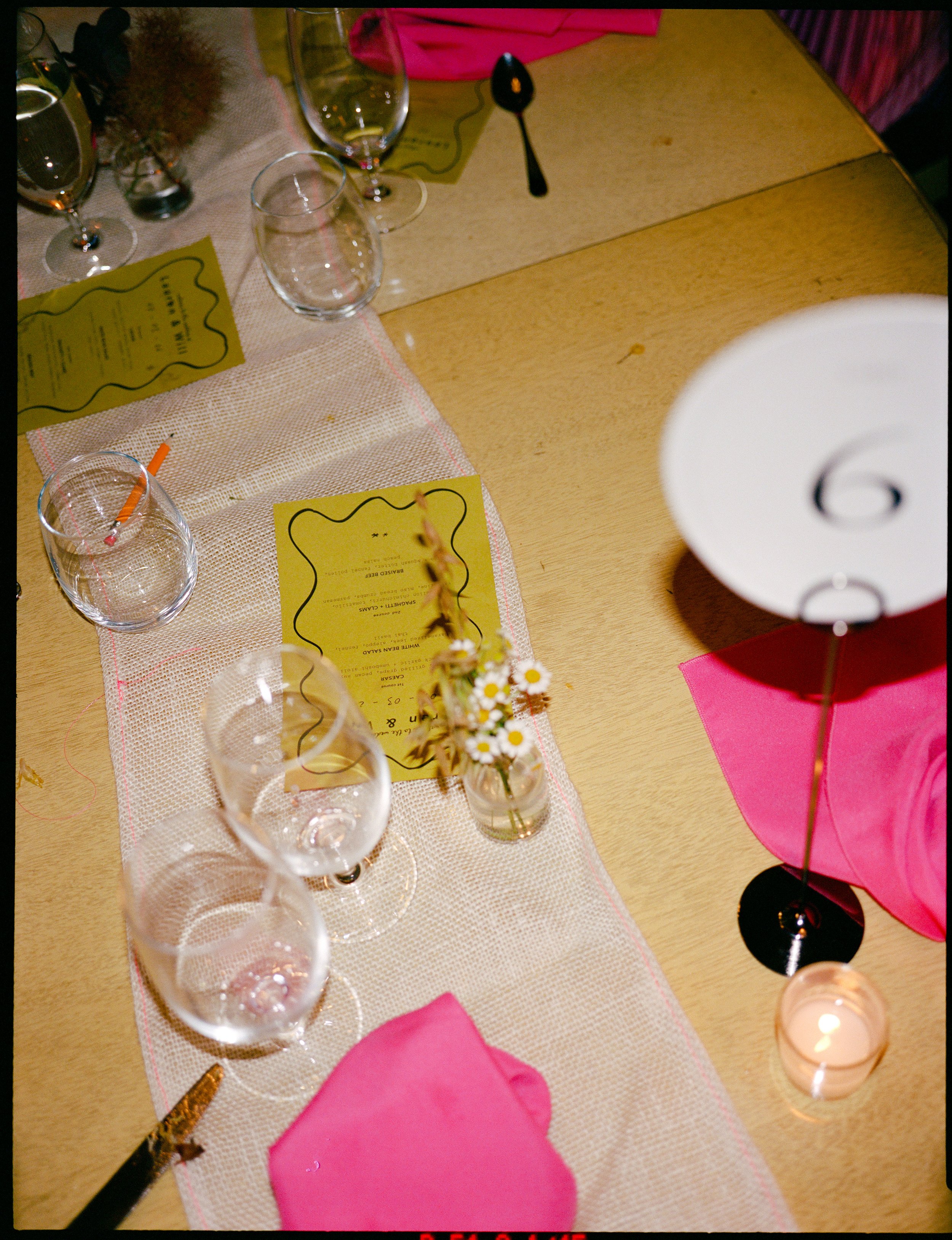 Empty wine glasses on a table decorated with pink napkins, a small flower arrangement with white daisies, candles, and a yellow menu in the background.