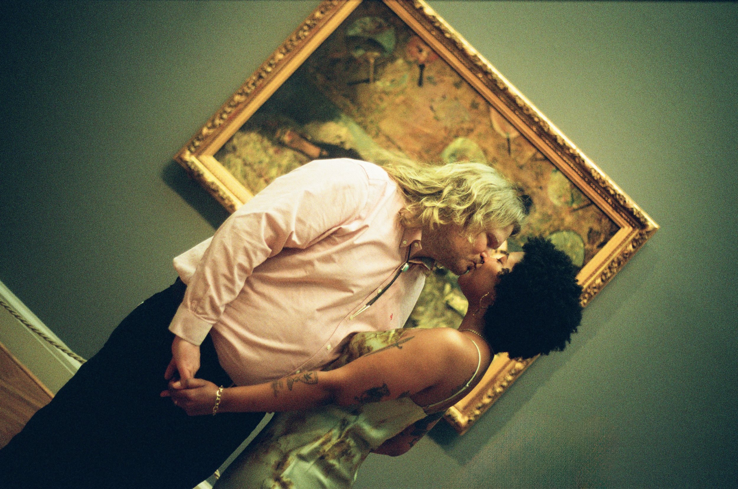 Two women sharing an intimate kiss in front of a framed painting on a green wall. One woman has curly blonde hair and is wearing a light pink shirt, while the other has dark curly hair and tattoos on her arm.
