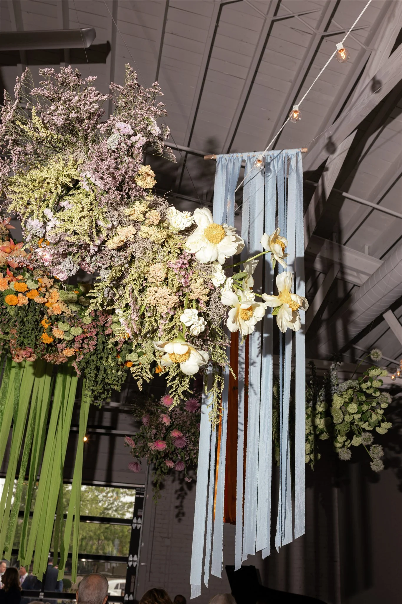 Large floral arrangement featuring various flowers including white peonies and pink, purple, and green blooms, displayed in an indoor setting with a ceiling decorated with hanging string lights and white ribbon strips.