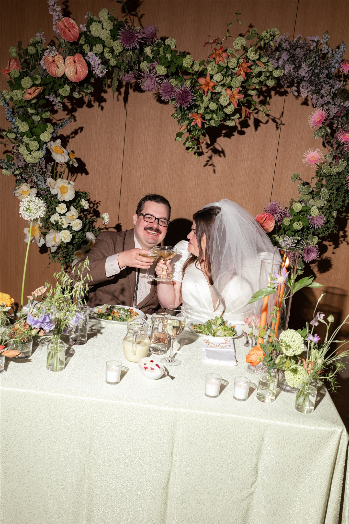 A bride and groom enjoying a toast at their wedding reception, seated at a table decorated with flowers and candles, with a floral arch overhead.