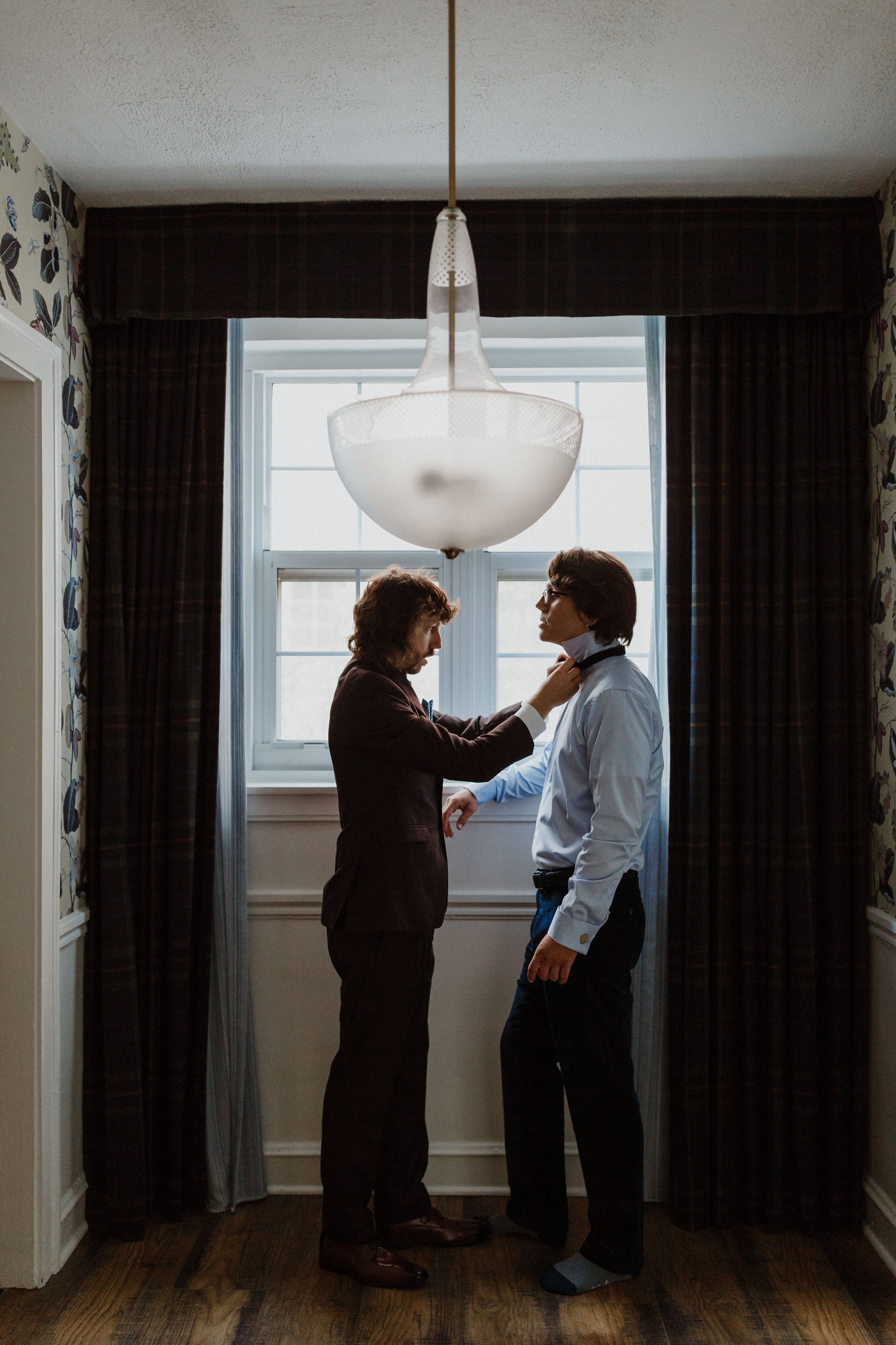 Two men, one adjusting the other's tie, standing face-to-face by a window with curtains, in a room with patterned wallpaper and wooden floor.