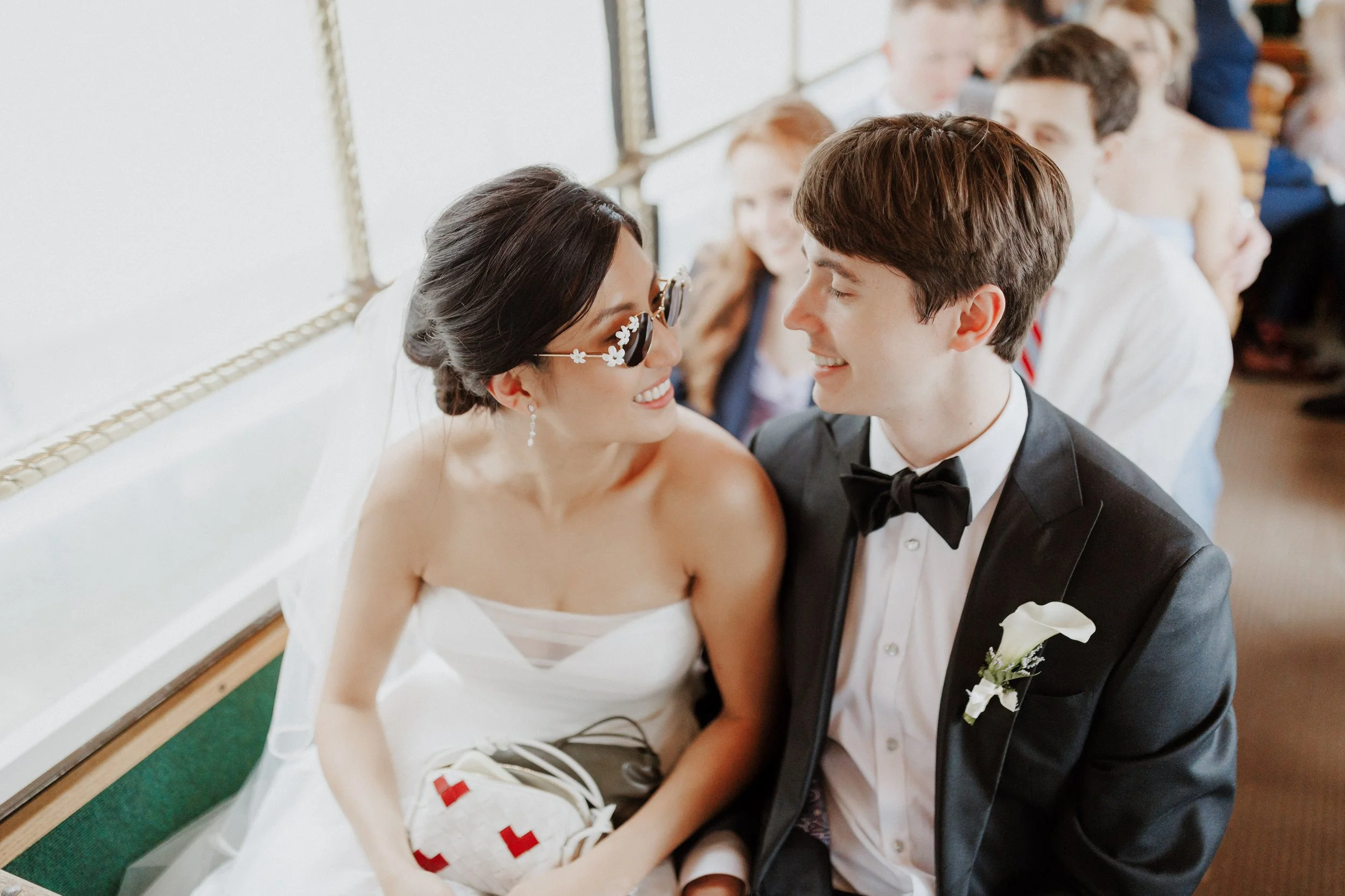 A bride and groom sitting closely and smiling at each other during their wedding ceremony in a church, with wedding guests in the background.