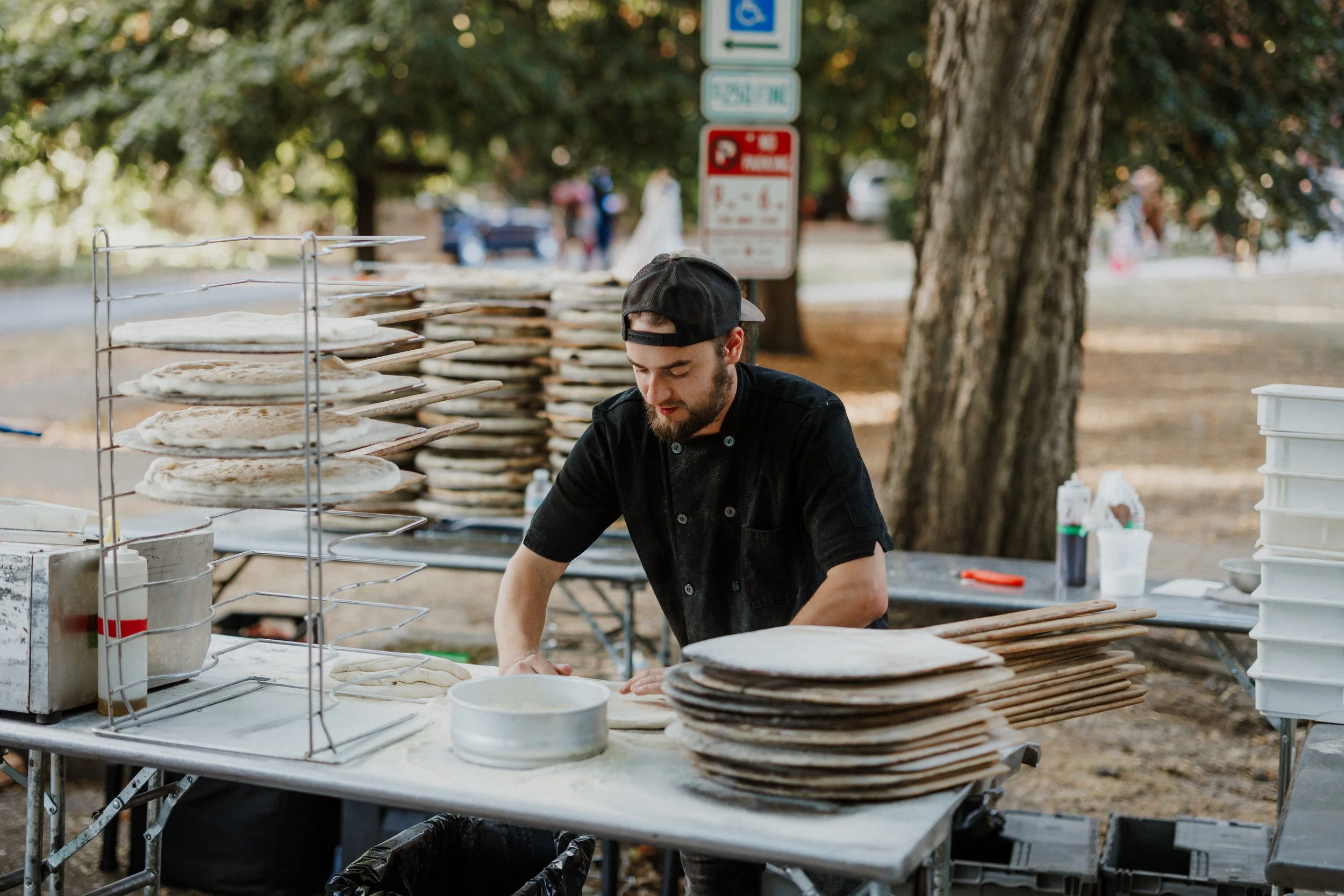 A man wearing a black chef coat and cap preparing pizza dough at an outdoor table. There are stacks of rolled pizza dough on metal shelves and various kitchen supplies around him. In the background, there are trees and traffic signs.