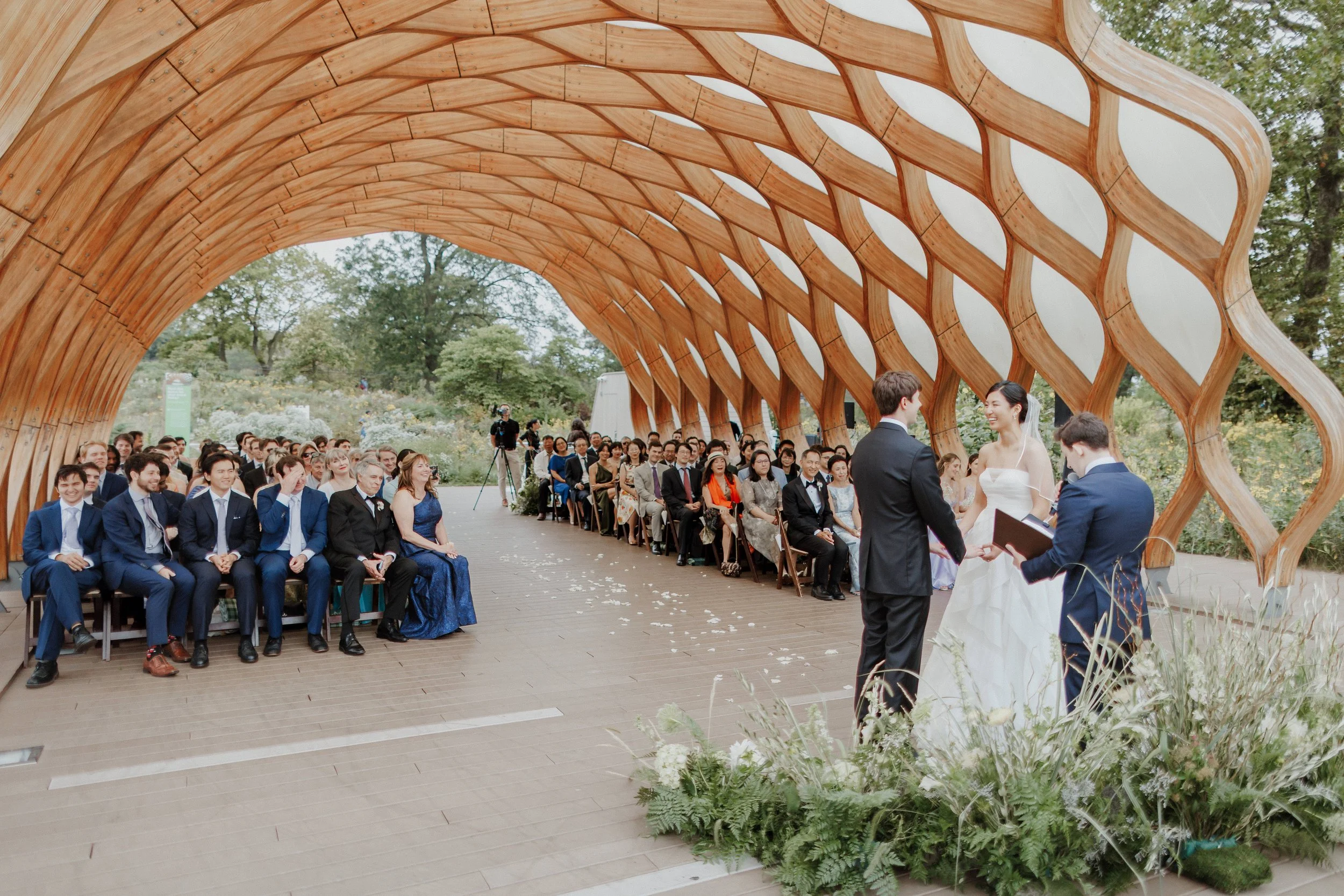 A wedding ceremony taking place under a curved wooden arch with guests sitting on both sides. The bride and groom are exchanging vows, and the officiant is reading from a book. The setting is outdoors with trees and lush greenery in the background.