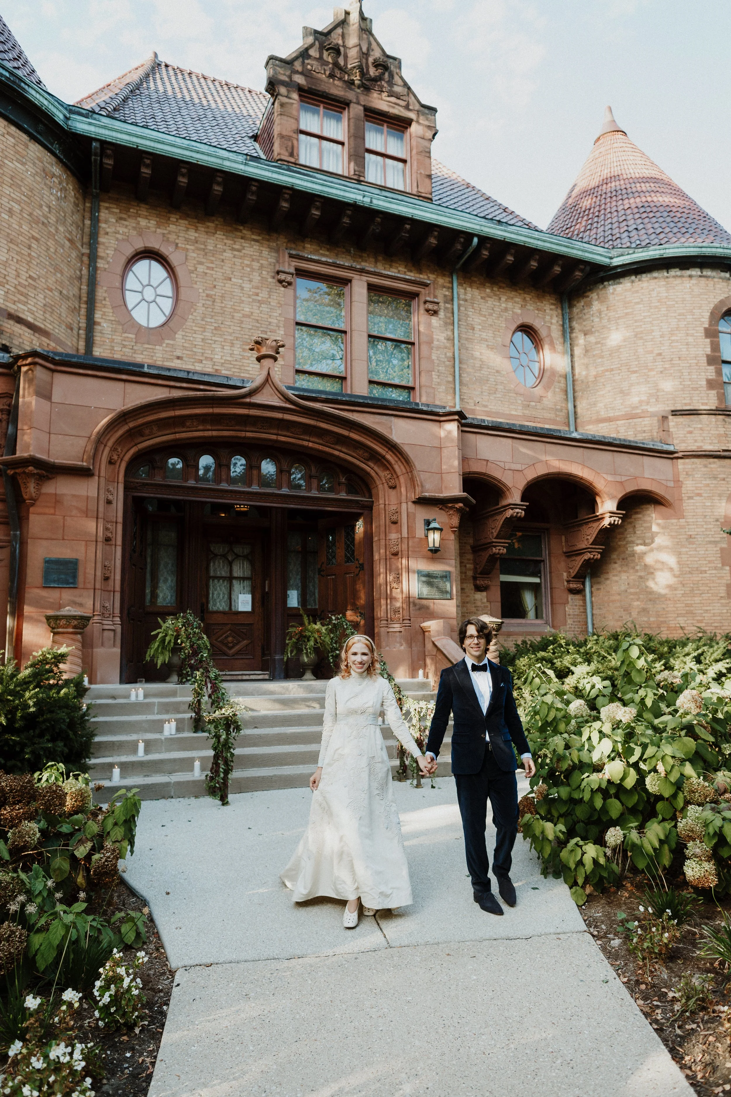 A bride and groom walking hand in hand down a pathway in front of a historic brick mansion. The bride is wearing a white wedding gown, and the groom is in a black tuxedo. The mansion has ornate architecture with a large wooden door, and the pathway i
