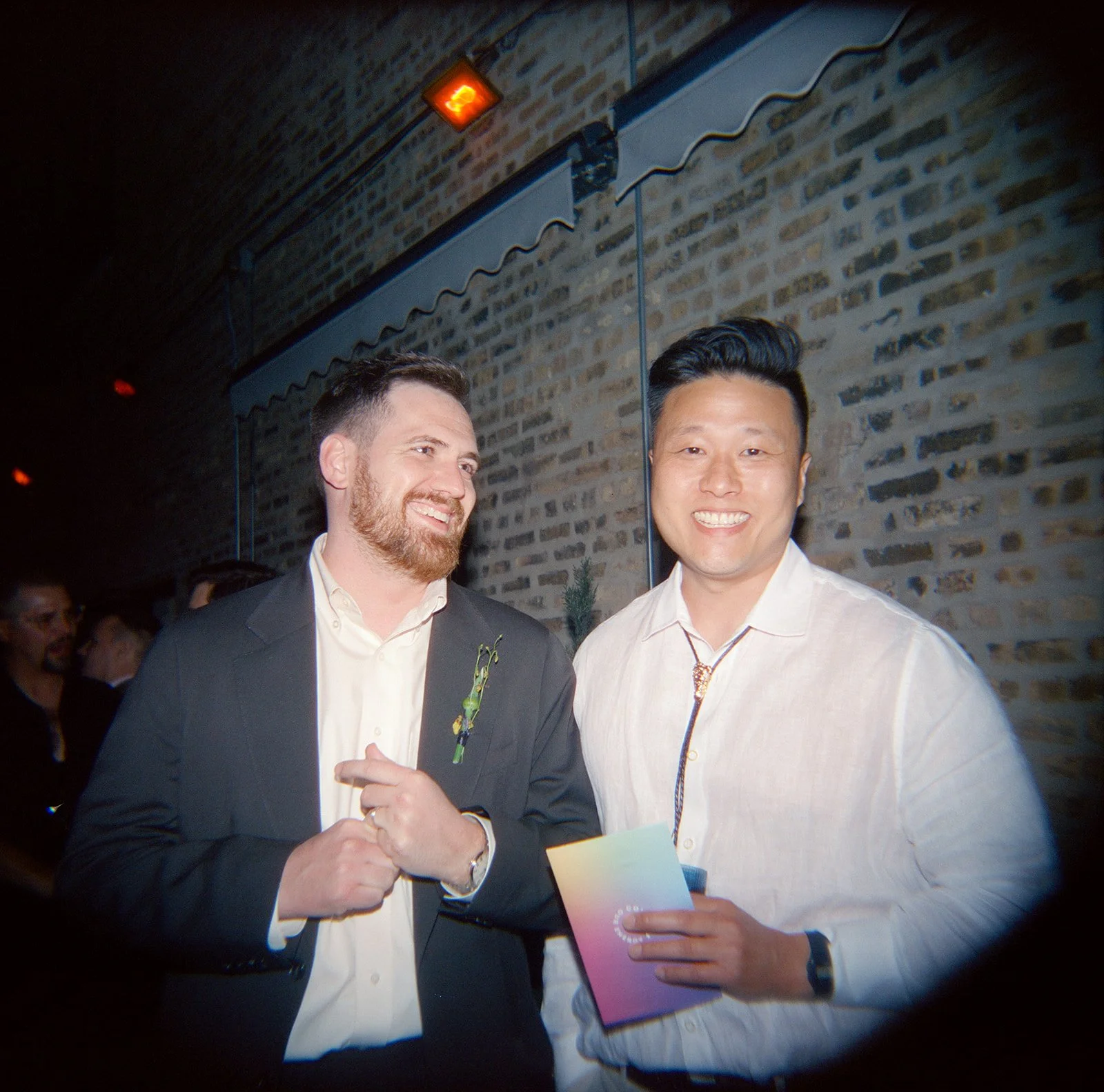 Two men smiling at an event, one wearing a suit and the other in a white shirt, holding a colorful brochure, with a brick wall and some people in the background.