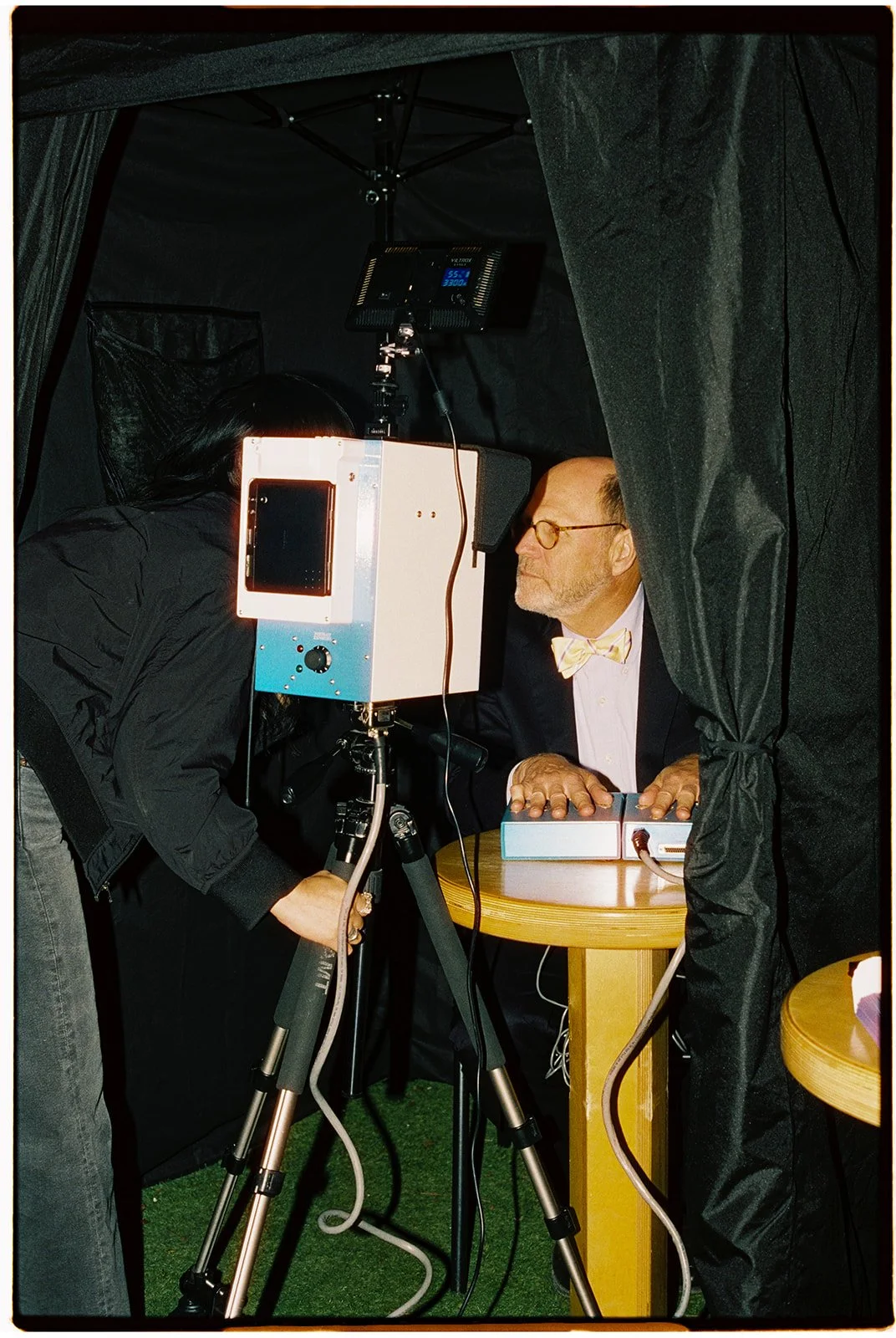 A man wearing glasses and a bow tie is under a black curtain, about to undergo an eye examination using a specialized medical device. A woman is adjusting the equipment near him.