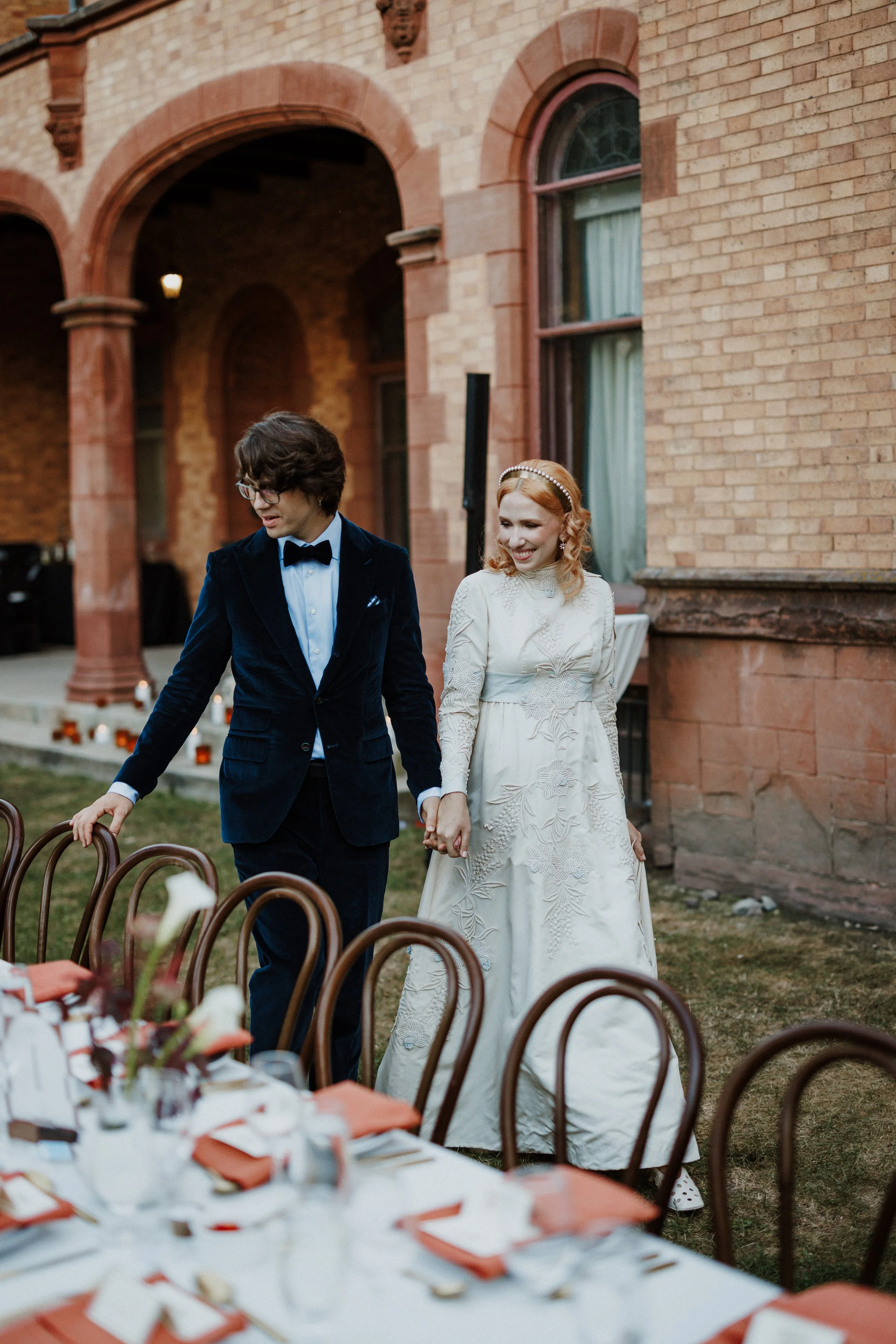 A bride and groom holding hands and smiling during an outdoor wedding reception, with a long table set with plates, napkins, and flowers in the foreground, in front of a brick building with arched windows.