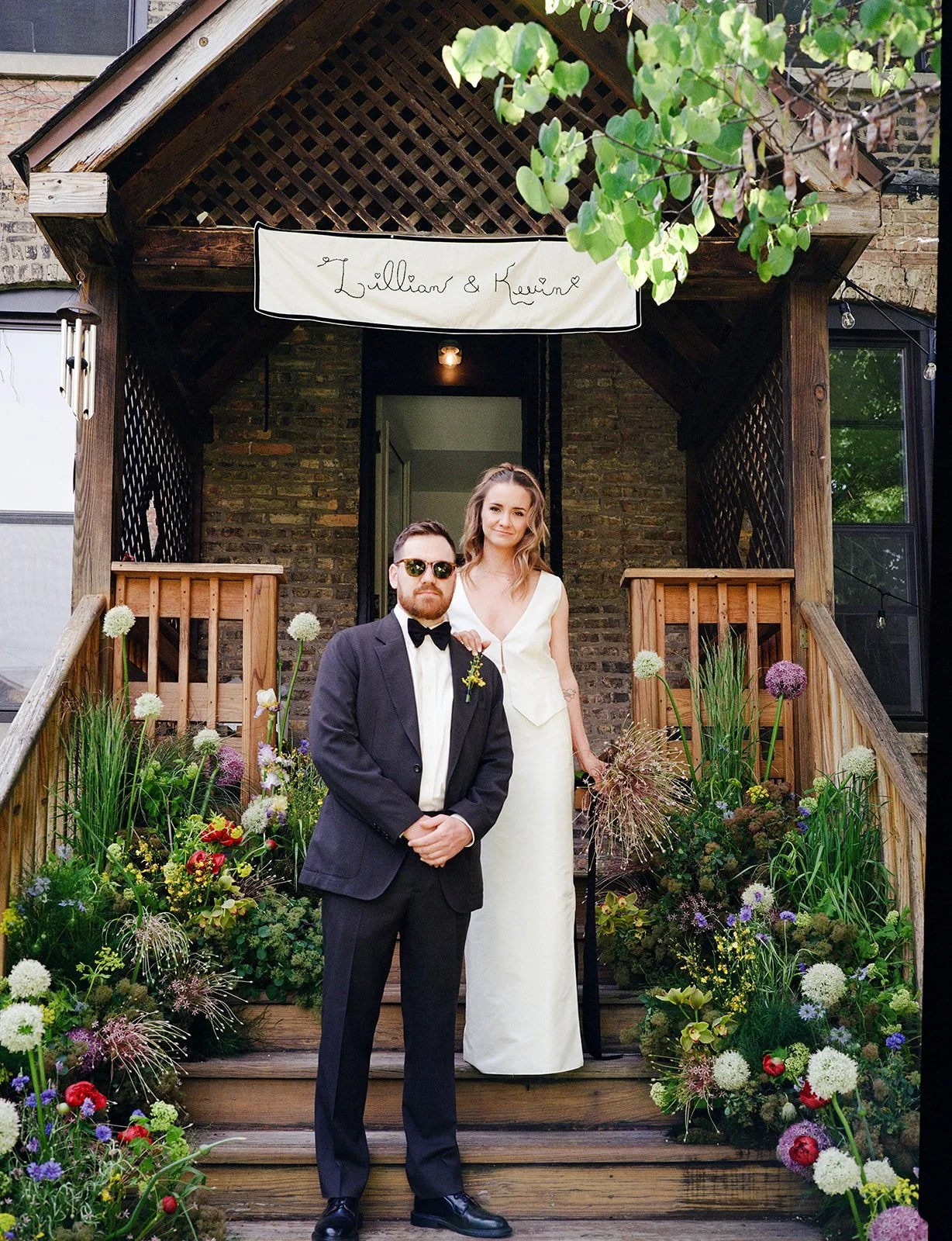 A man in a tuxedo with sunglasses and a woman in a white dress standing on a wooden porch decorated with colorful flowers, with a sign above that reads 'Lillian & Kevin'.