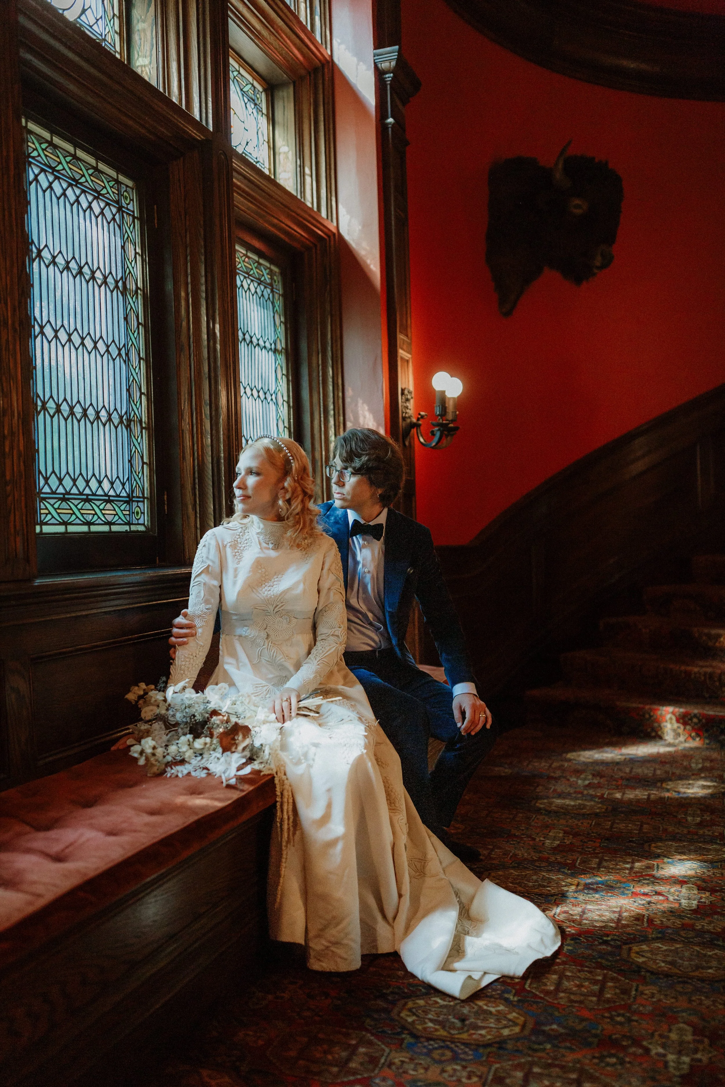 A bride and groom sitting in a vintage, wood-paneled room with stained glass windows. The bride, wearing a white lace wedding gown, holds a bouquet of flowers, while the groom, dressed in a tuxedo, sits close beside her. A mounted buffalo head is seen on the red wall behind them, and a rug covers the wooden floor.