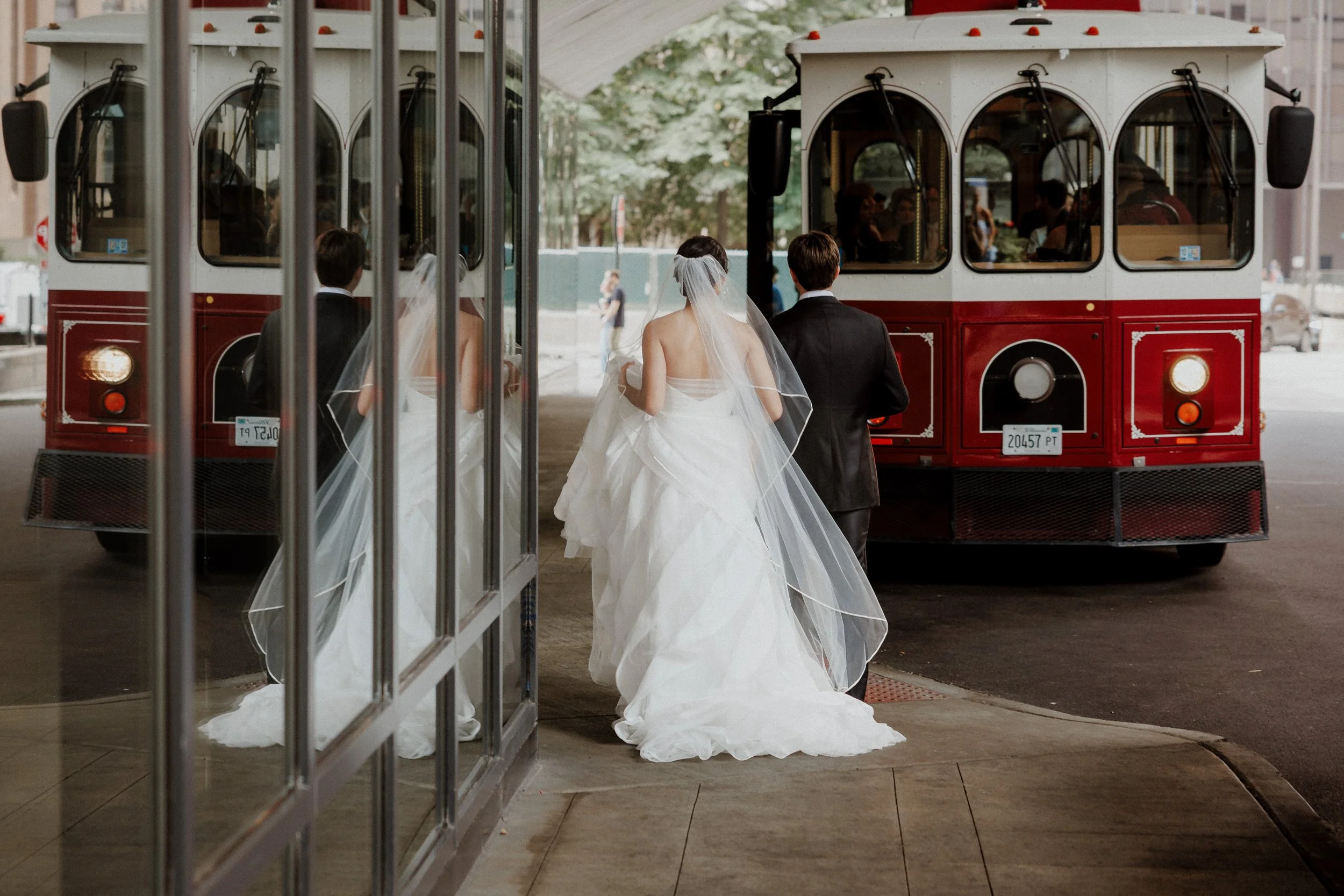A bride in a white wedding gown and veil walking out of a tram, accompanied by a groom in a black suit. Reflection of the couple and other wedding guests can be seen in the tram windows.