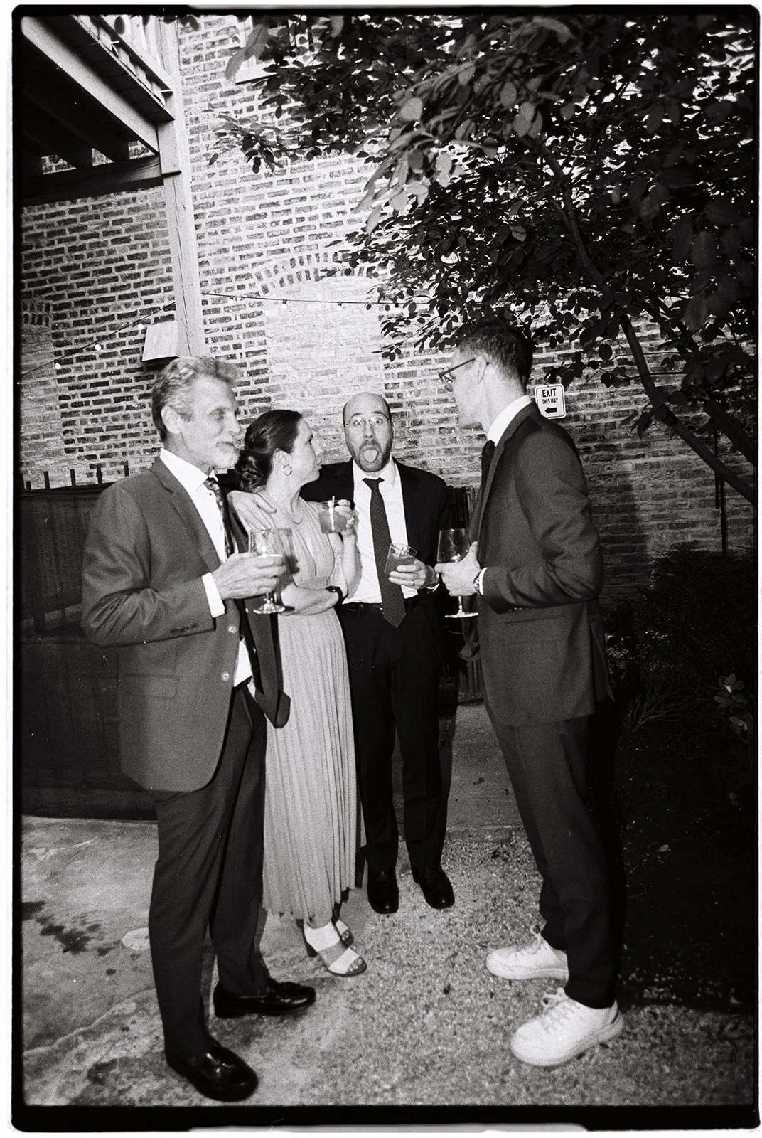 Four people in formal attire socializing outdoors, holding drinks, near brick walls and trees, at night.