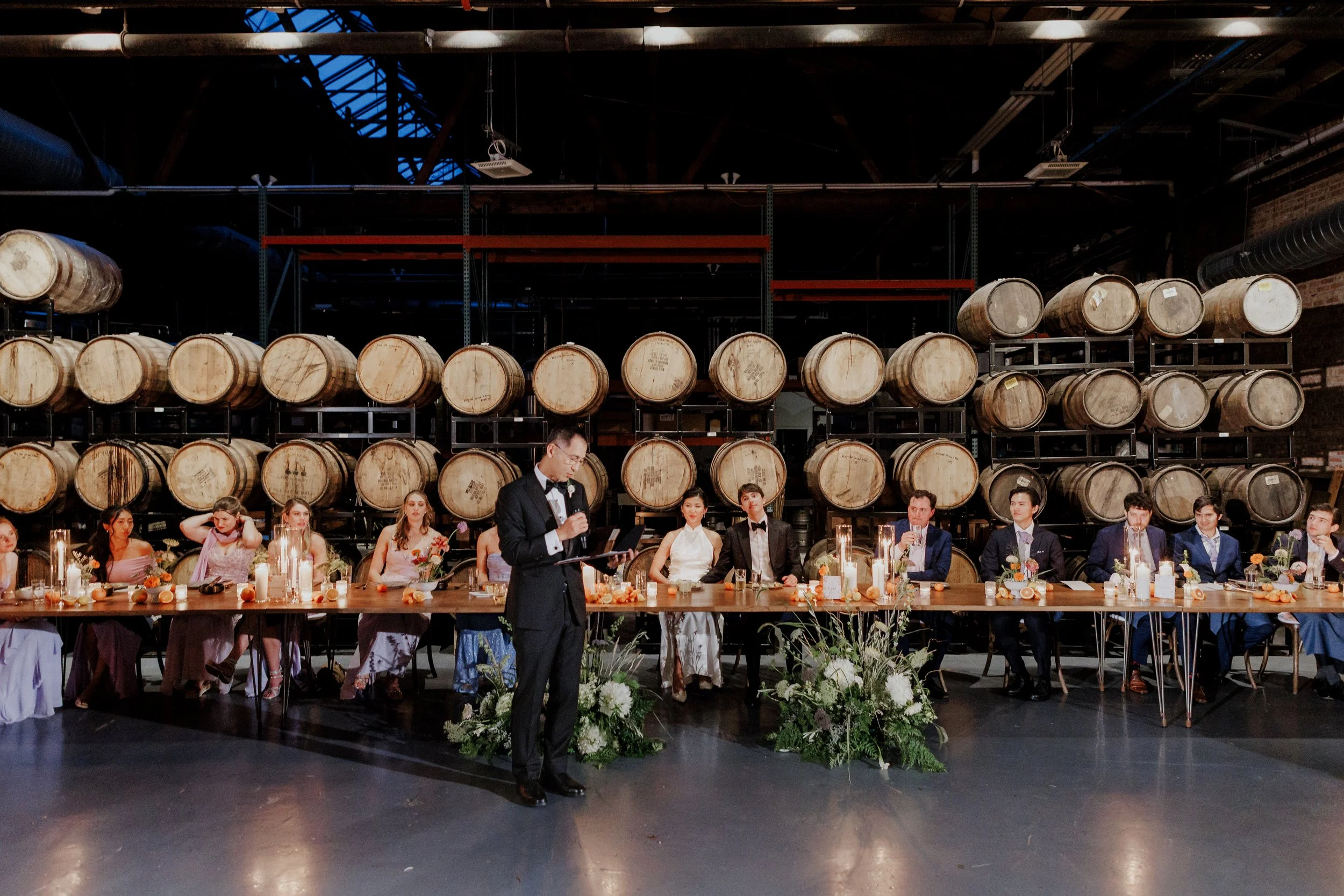 A wedding reception with the bride and groom seated at a long table decorated with flowers and candles, surrounded by guests. The setting includes a backdrop of stacked barrels and industrial elements such as metal shelving and exposed pipes and bric