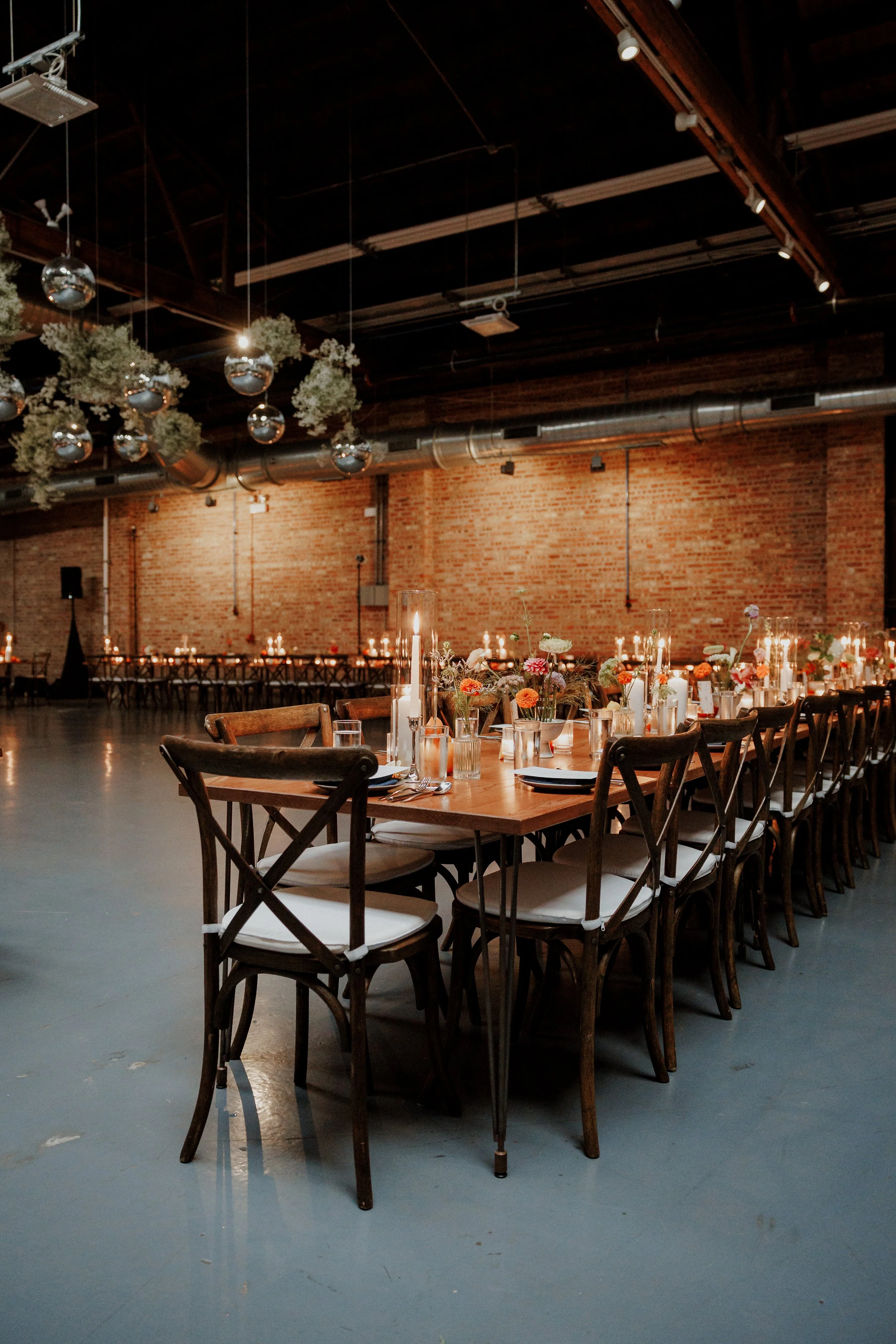 Wedding reception setup with long tables decorated with candles, flowers, and tall candles, inside an industrial-style venue with brick walls, a dark ceiling, and hanging greenery and reflective ornaments.
