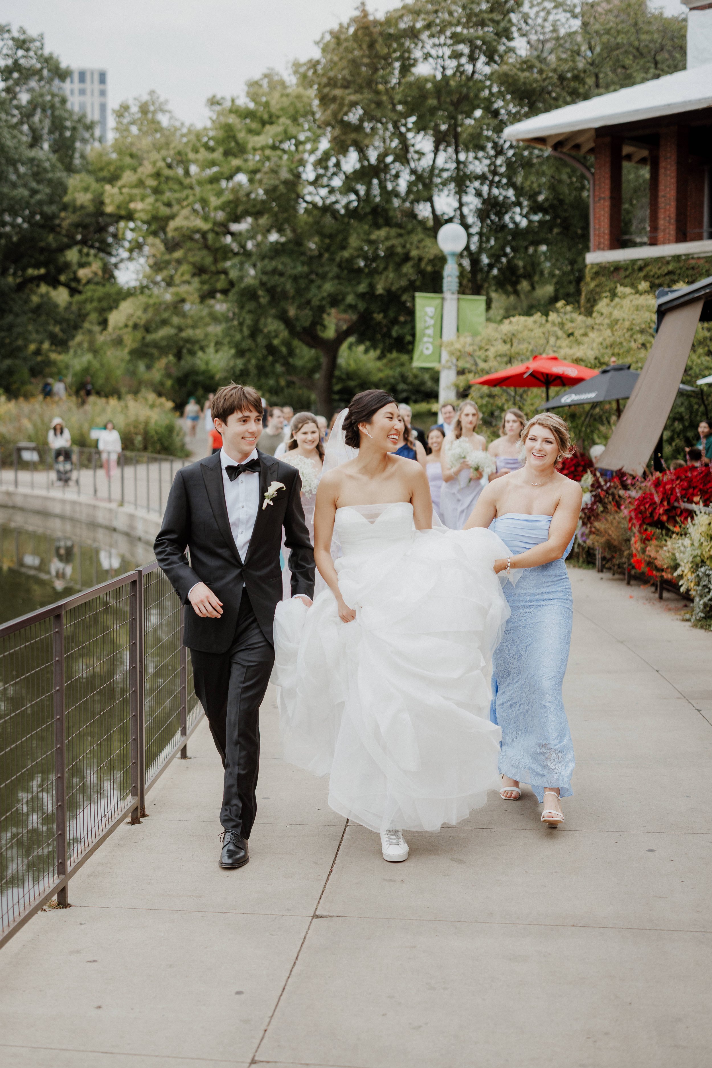 Bride and groom walking together outdoors, smiling, with bridesmaids in the background, near water with a railing, city buildings, trees, and outdoor umbrellas.