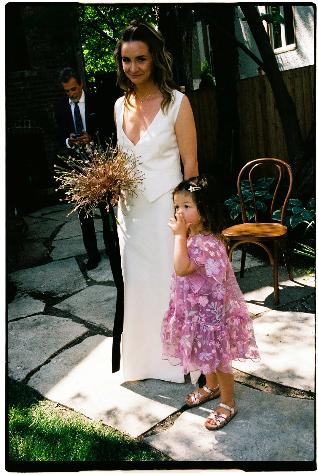 A woman in a white dress holding a bouquet of flowers outdoors, with a young girl in a pink dress standing next to her, covering her mouth. A man in a suit is in the background using a phone.