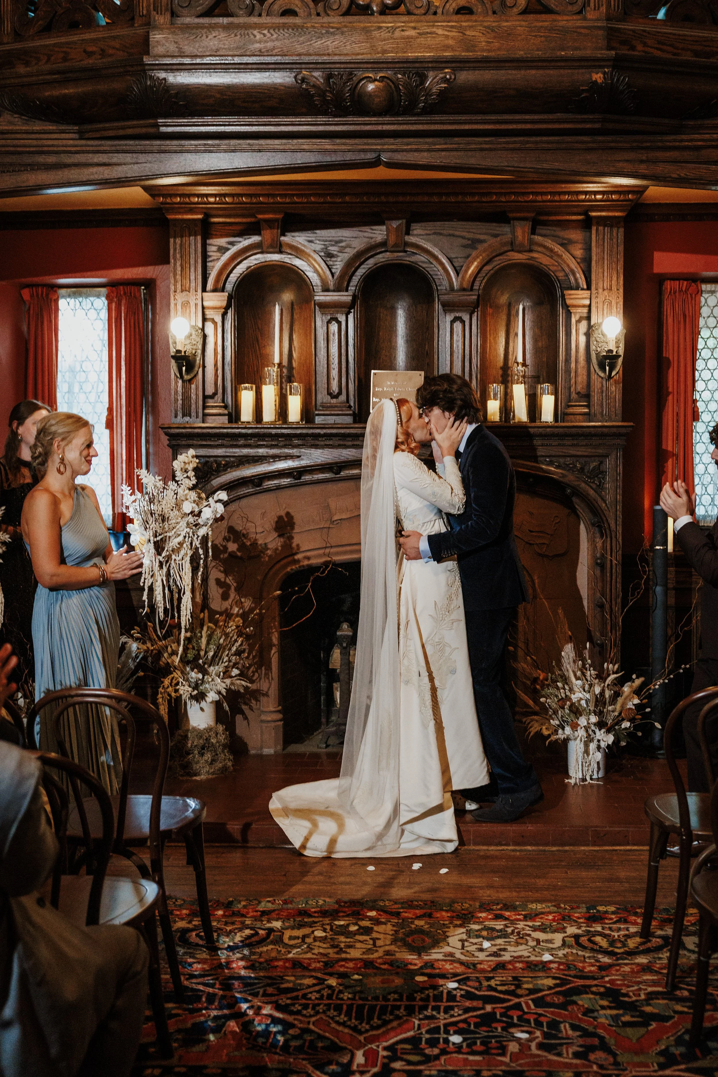 A wedding ceremony with a bride and groom kissing in front of a fireplace, surrounded by guests, candles, flowers, and ornate woodwork in a decorated room.